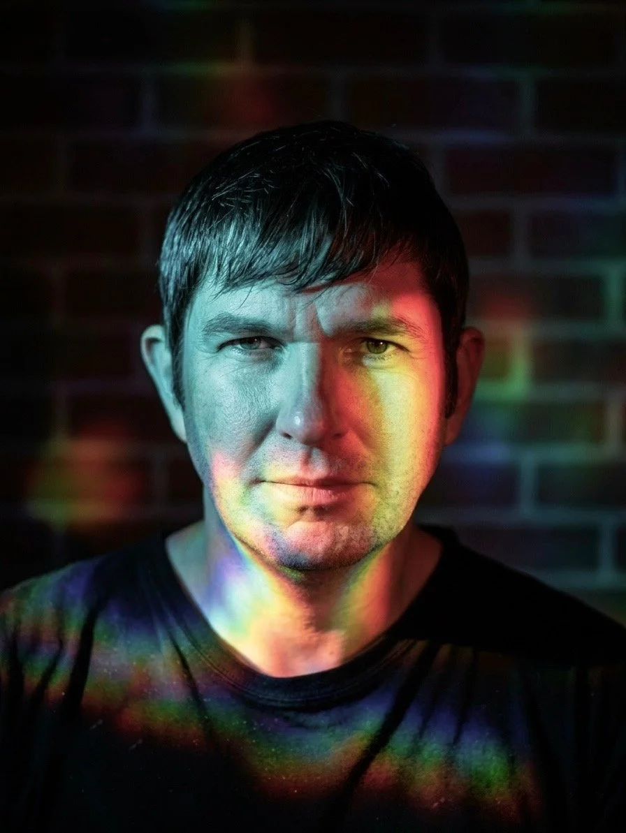 A man with short brown hair looking directly at the camera, illuminated by colorful rainbow light patches on his face and black shirt, with a blurred brick wall background.