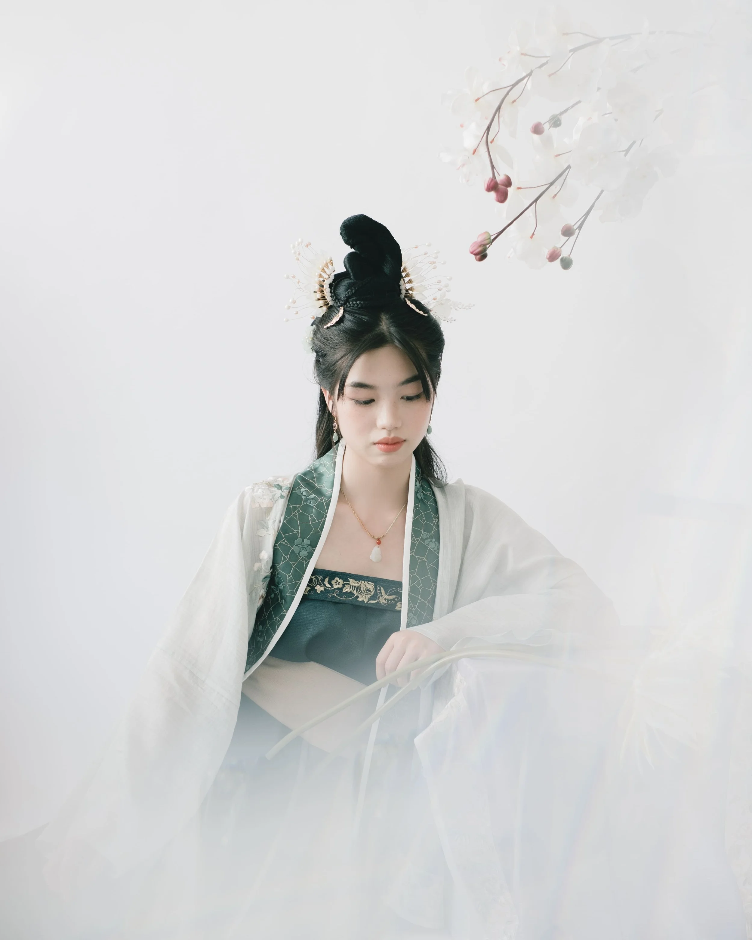 A young woman dressed in traditional East Asian clothing, with an intricately styled hairdo adorned with hairpieces and earrings, is looking down, possibly reading or examining something, against a plain white background, with a branch of white and p
