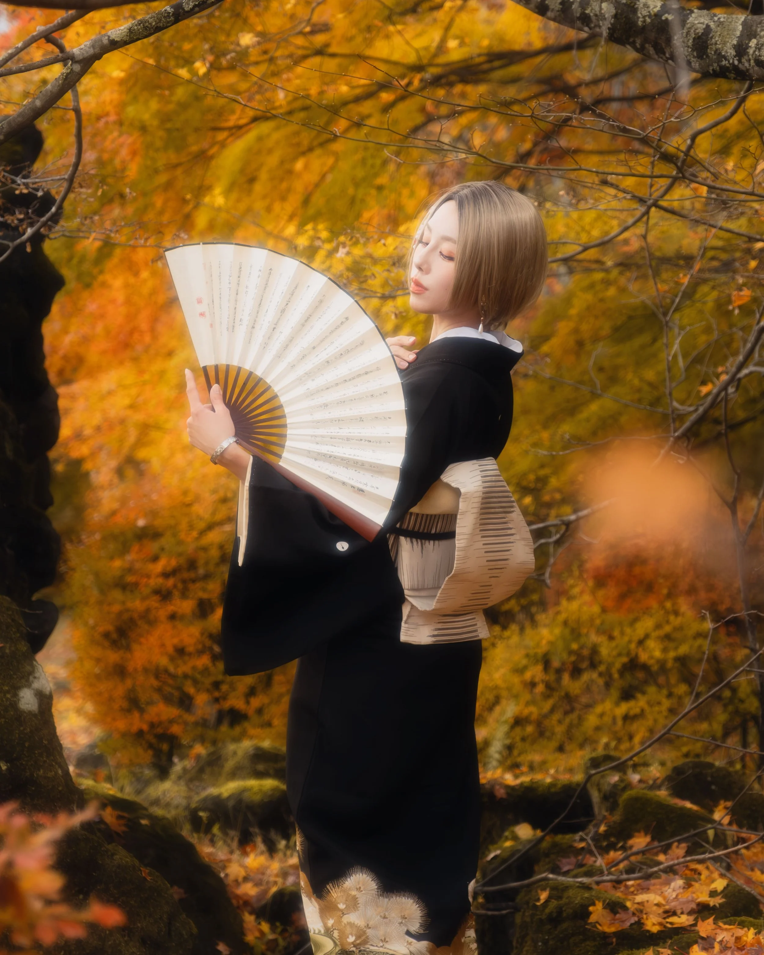 A woman standing outdoors in autumn, holding a traditional folding fan with Japanese characters, wearing a black kimono with gold and white patterns, amidst orange and yellow leaves on trees.