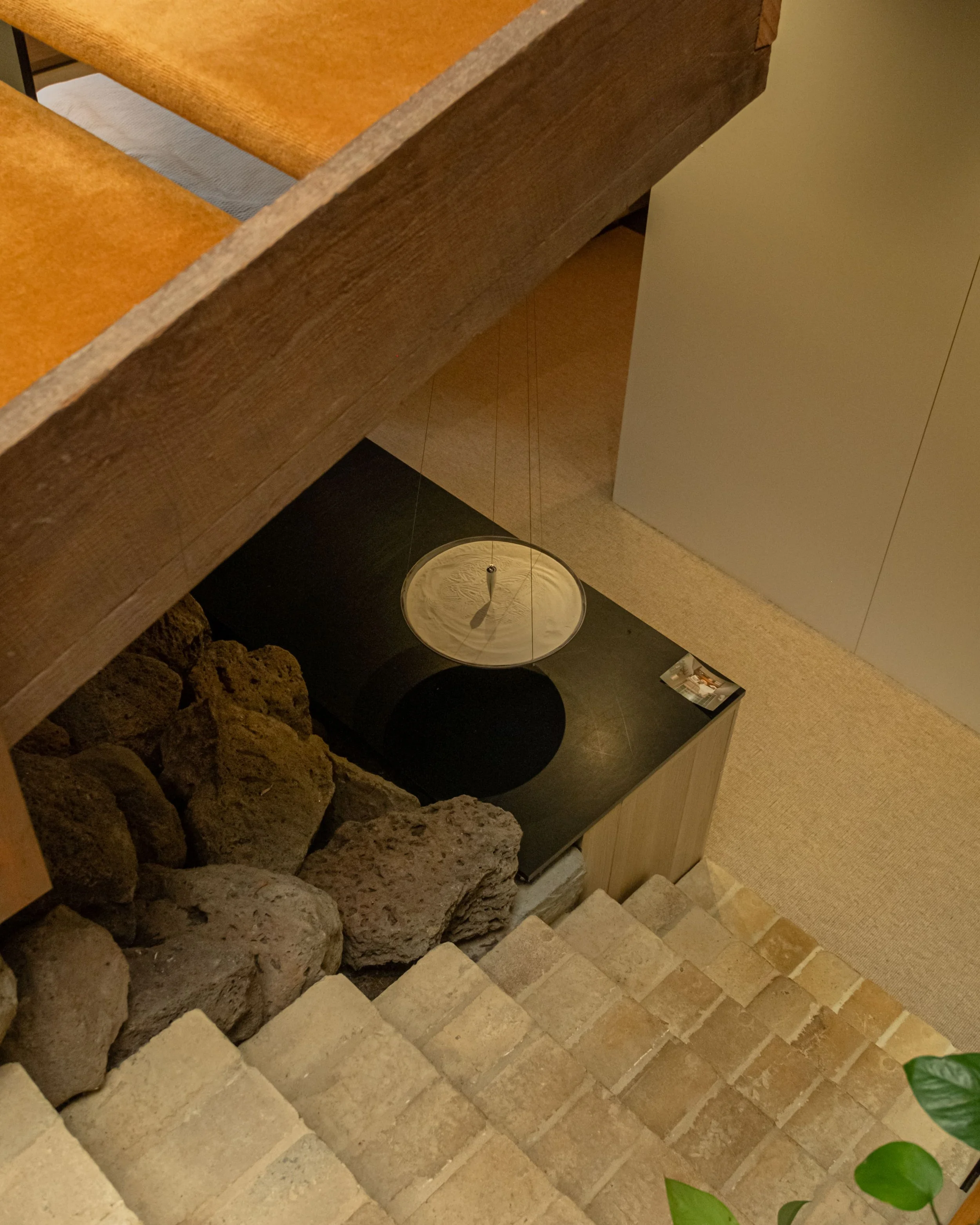 View looking down a staircase with brick steps, showing a black table or countertop, a white decorative hanging light, wooden beams, rocks, and a beige carpeted floor.