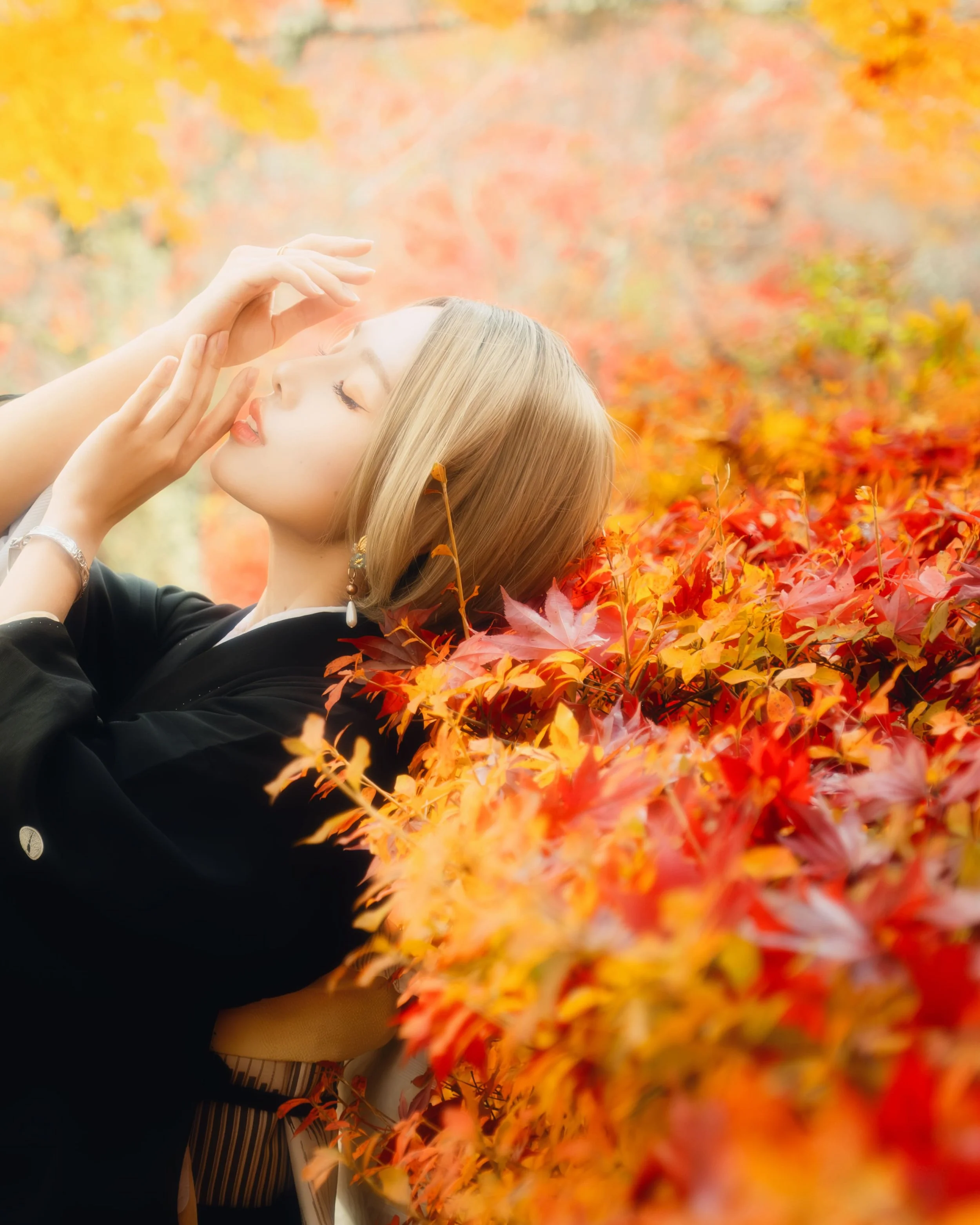 A young woman with blonde hair and earrings lying on a bed of colorful fall leaves with her eyes closed.