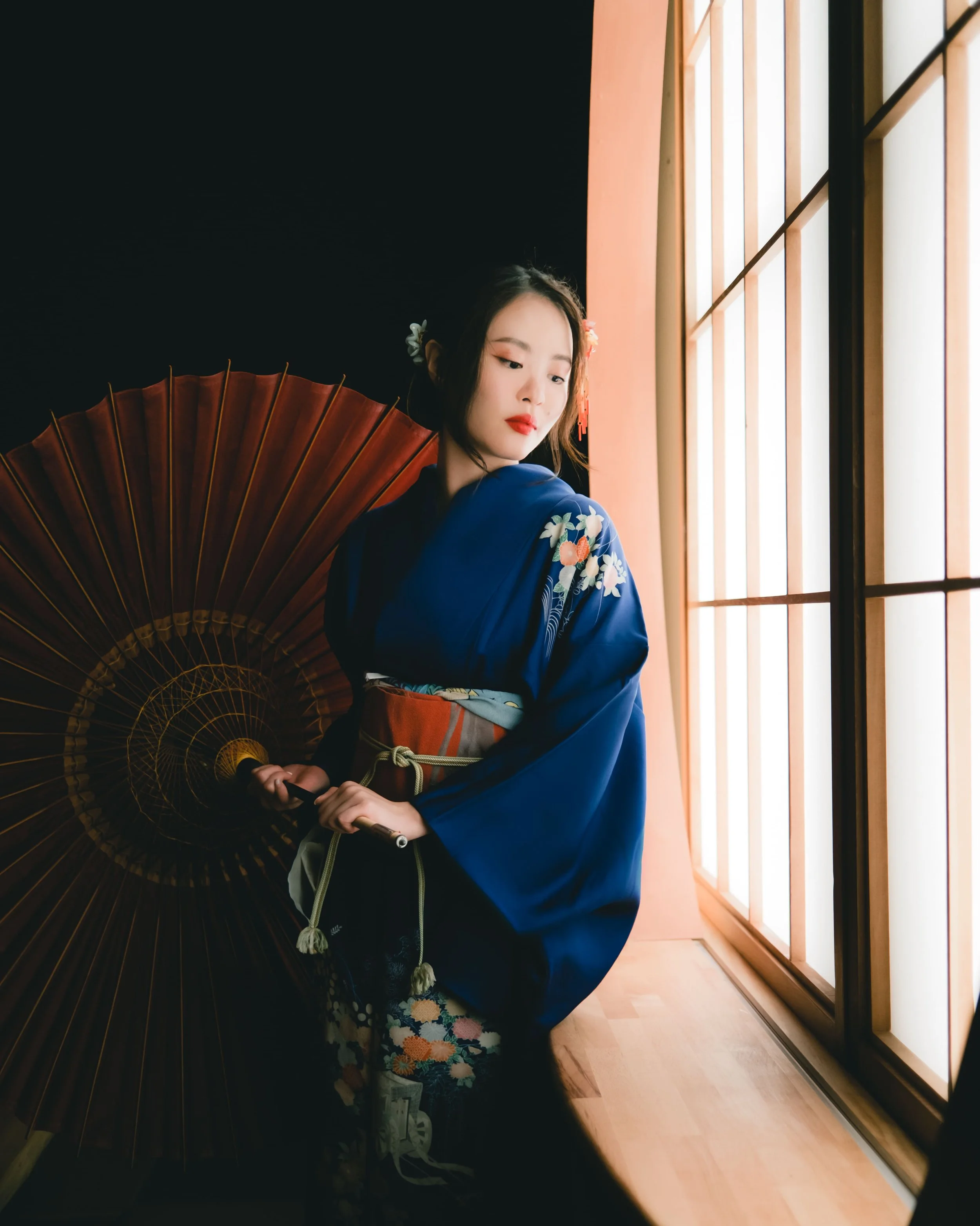 A young woman wearing a blue kimono with floral patterns is holding a traditional Japanese paper parasol. She is standing near a large window with wooden frames, and sunlight is illuminating her face.
