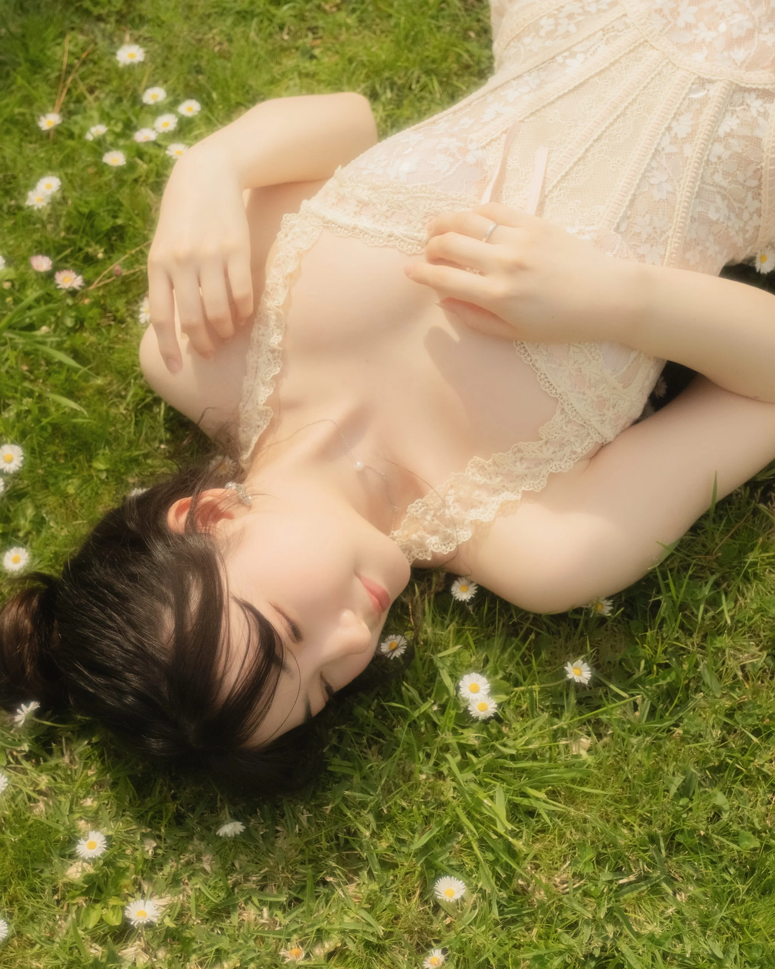 A woman lies on grass with small white daisies, wearing a lace top and jewelry, appearing relaxed and peaceful with her eyes closed.