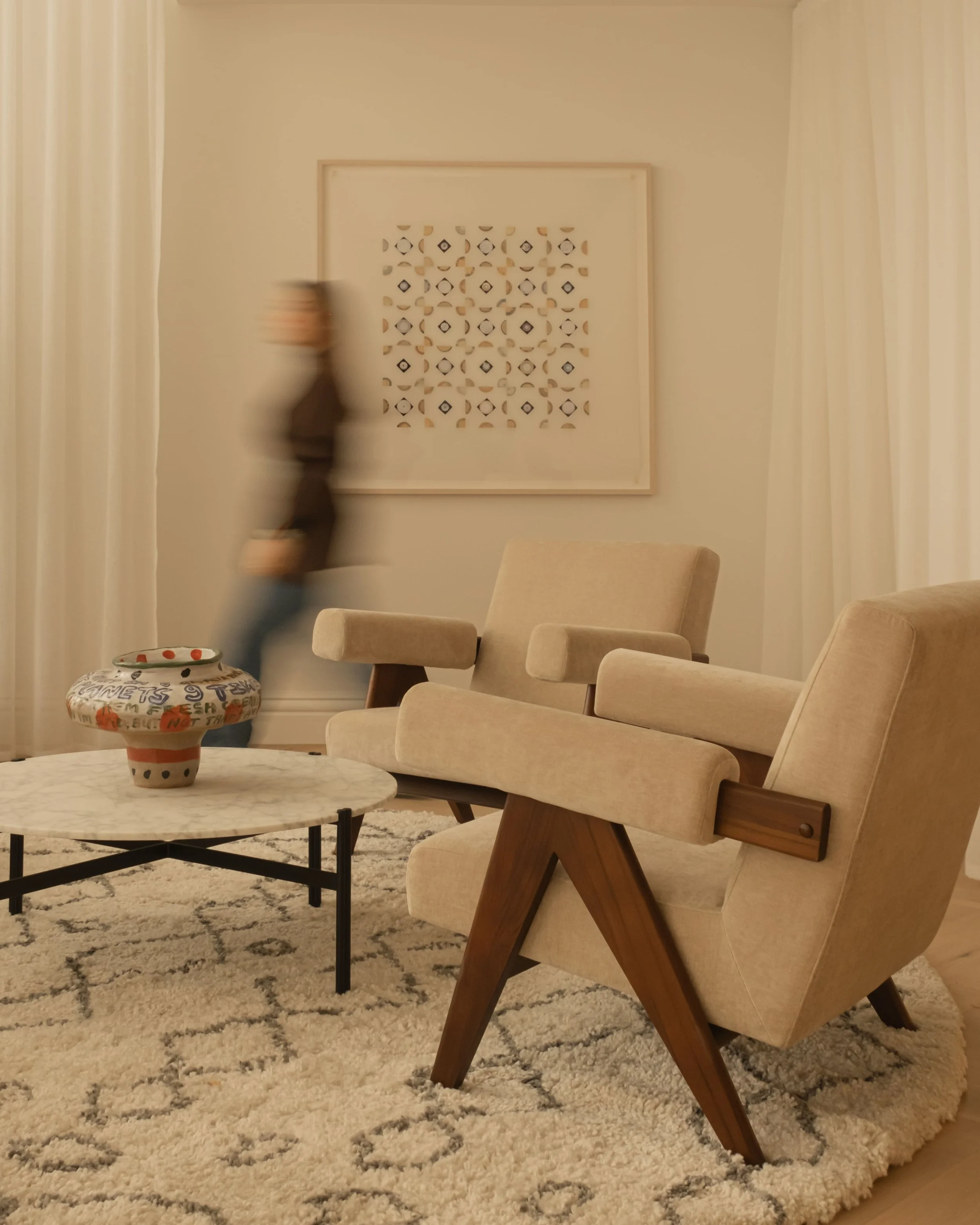 Living room with beige armchairs, a round marble coffee table with a decorative vase, a textured rug, and abstract wall art. A person is walking past in the background.