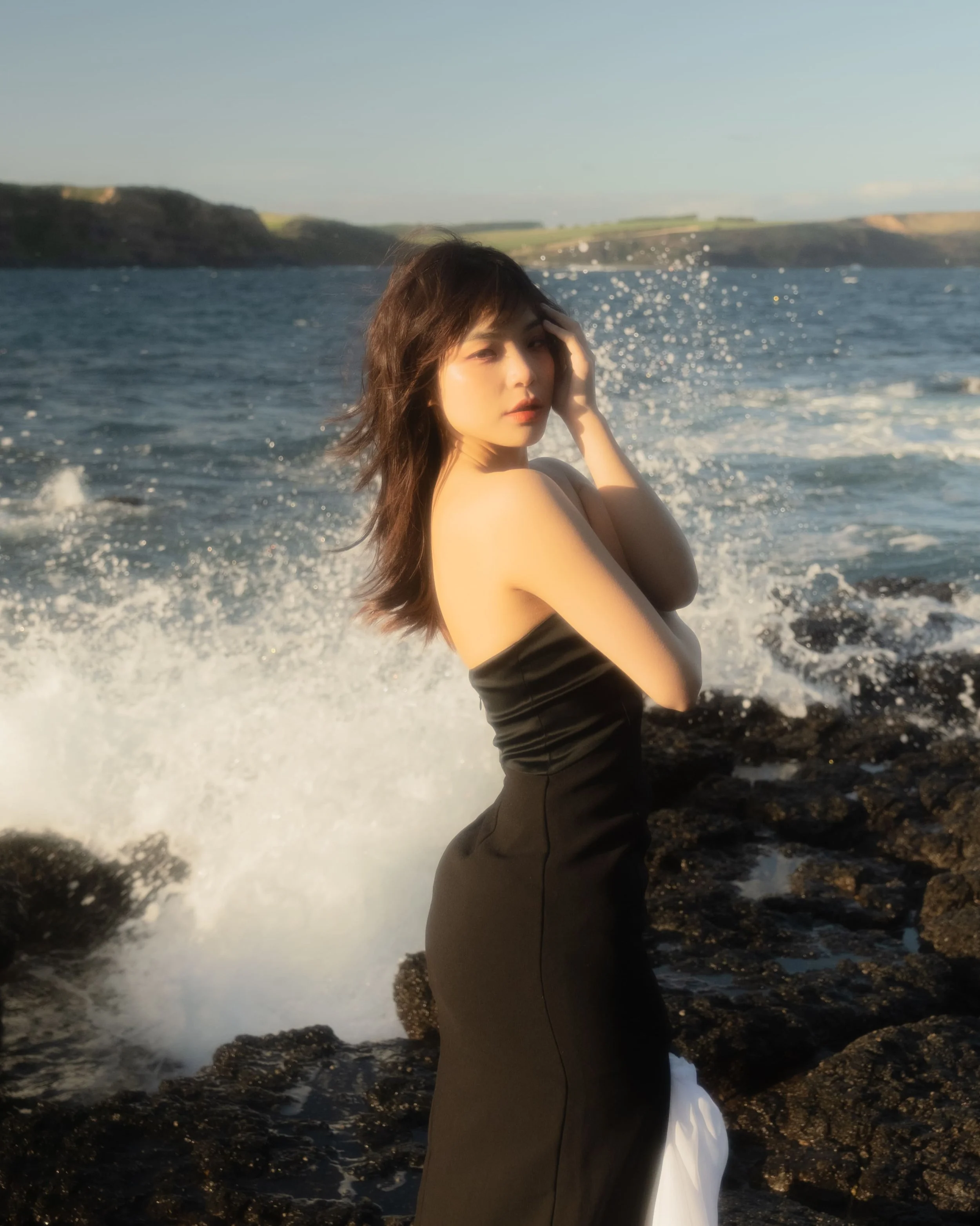 A woman in a black strapless dress standing on rocky shoreline with waves crashing behind her and ocean in the background.