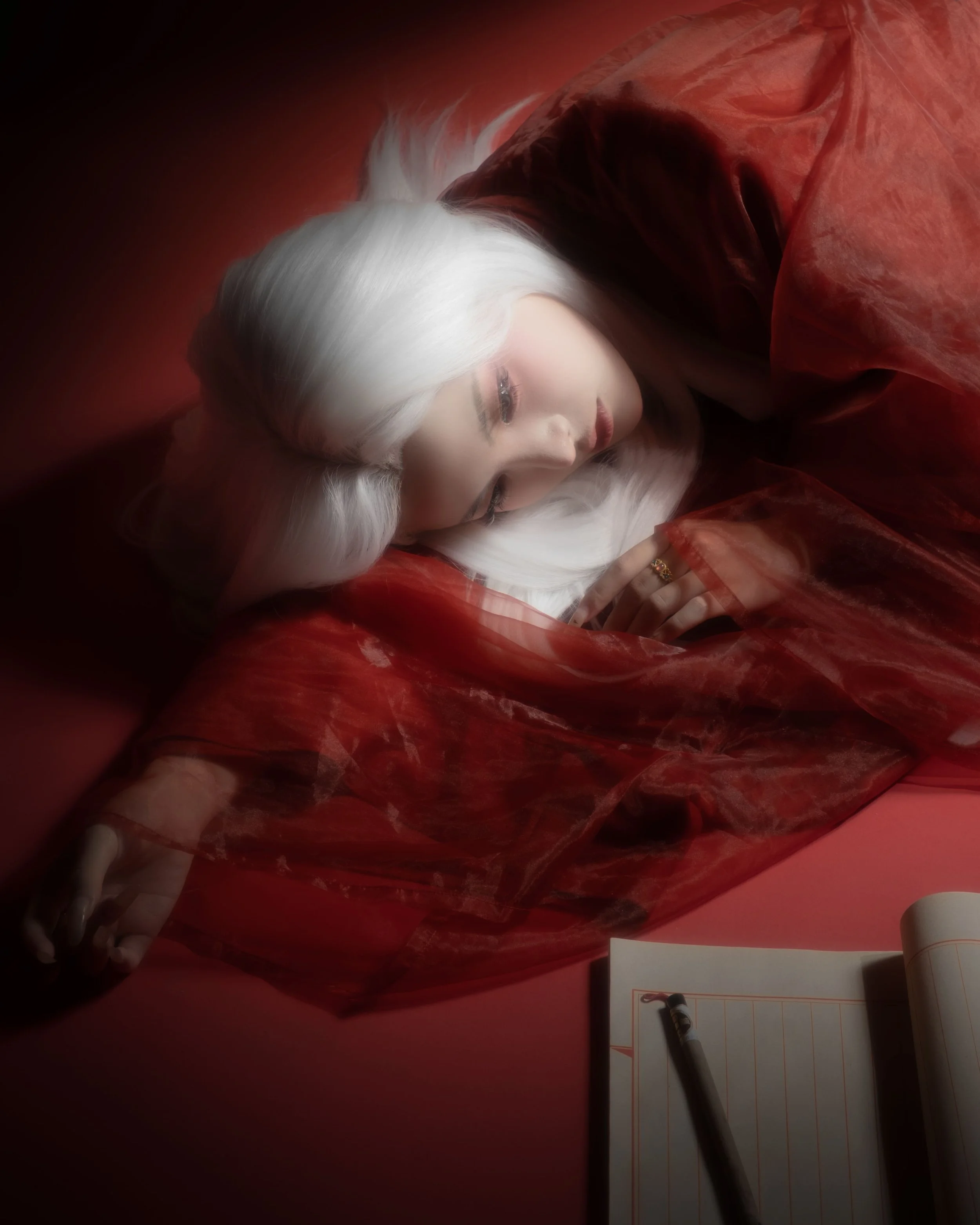 A woman with long platinum blonde hair lying on her side, wearing a red sheer jacket, resting on a red surface with a dark background. There is a notepad and pen next to her.