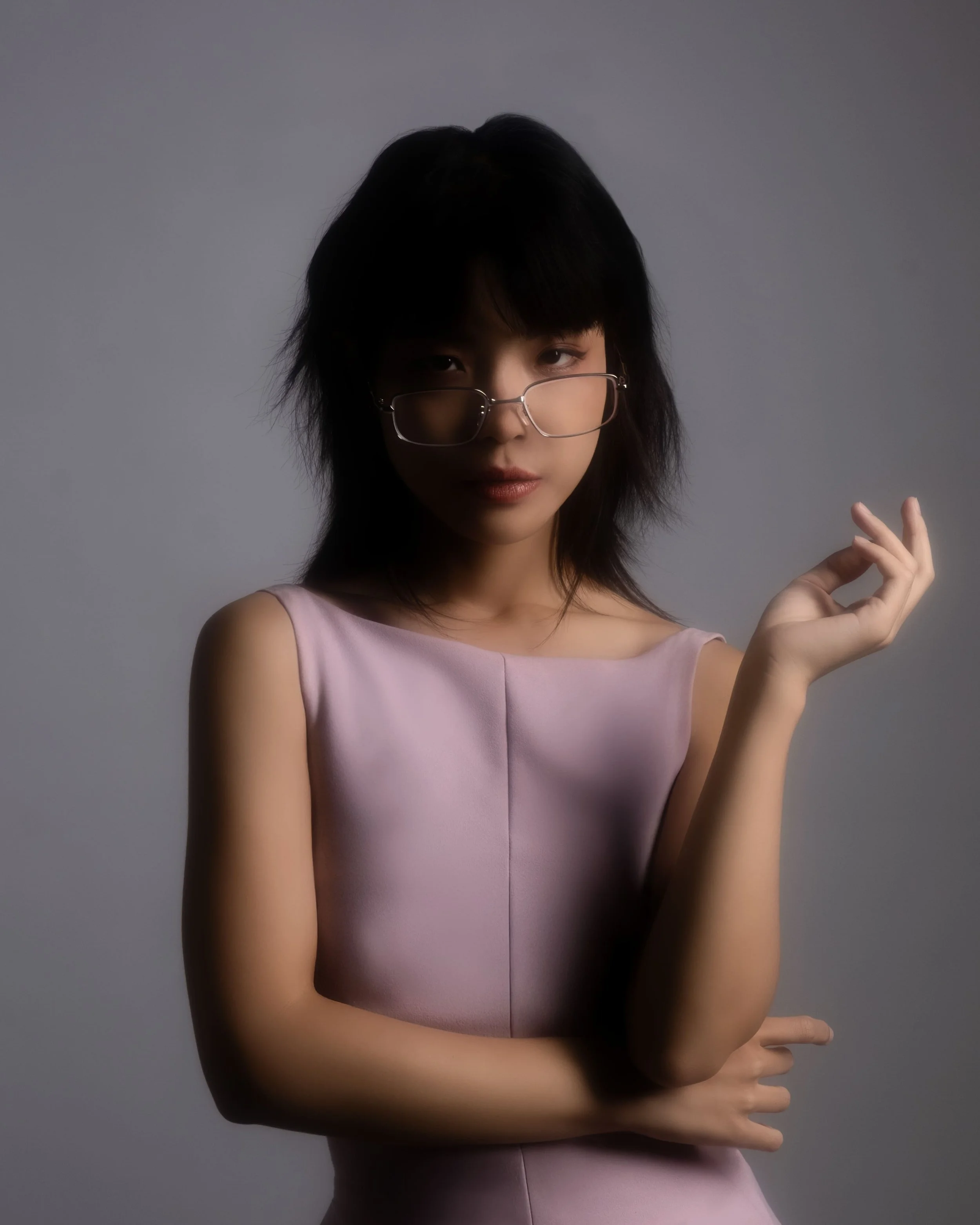 A woman with dark hair and glasses wearing a sleeveless lavender dress, posing against a plain gray background.