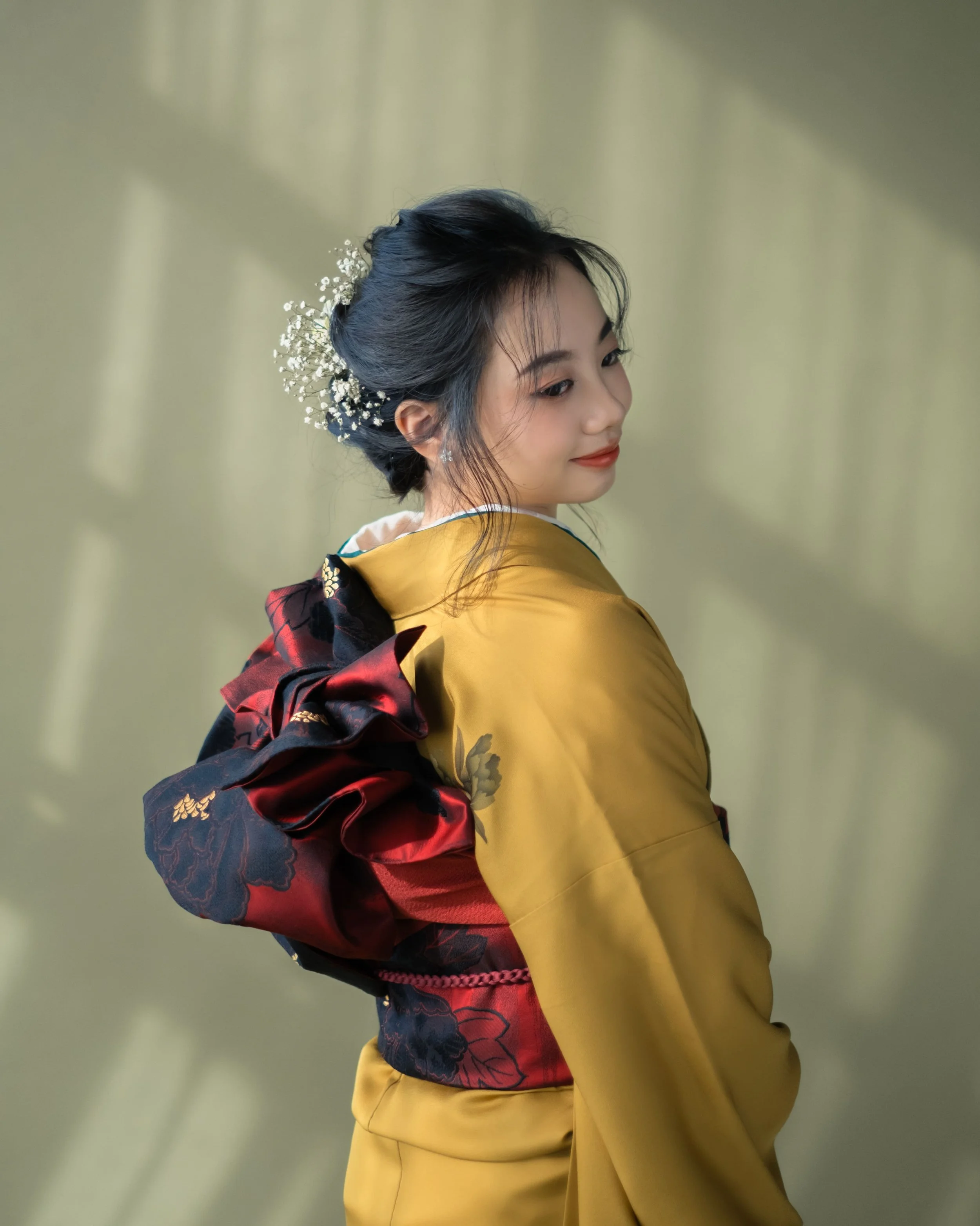 A woman dressed in traditional Japanese attire with her hair styled elegantly and adorned with white floral hair accessories, looking slightly downward and to her left, with a soft expression on her face.