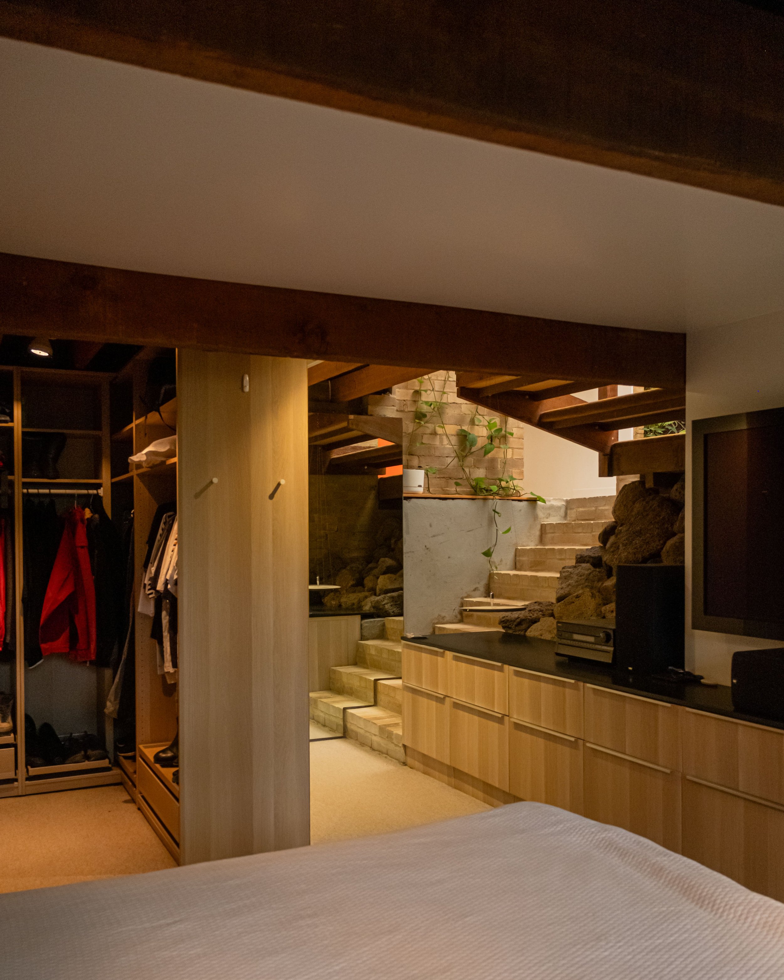 Interior view of a bedroom with a wooden closet, stairs, and natural rock decor behind a television, with a bed in the foreground.