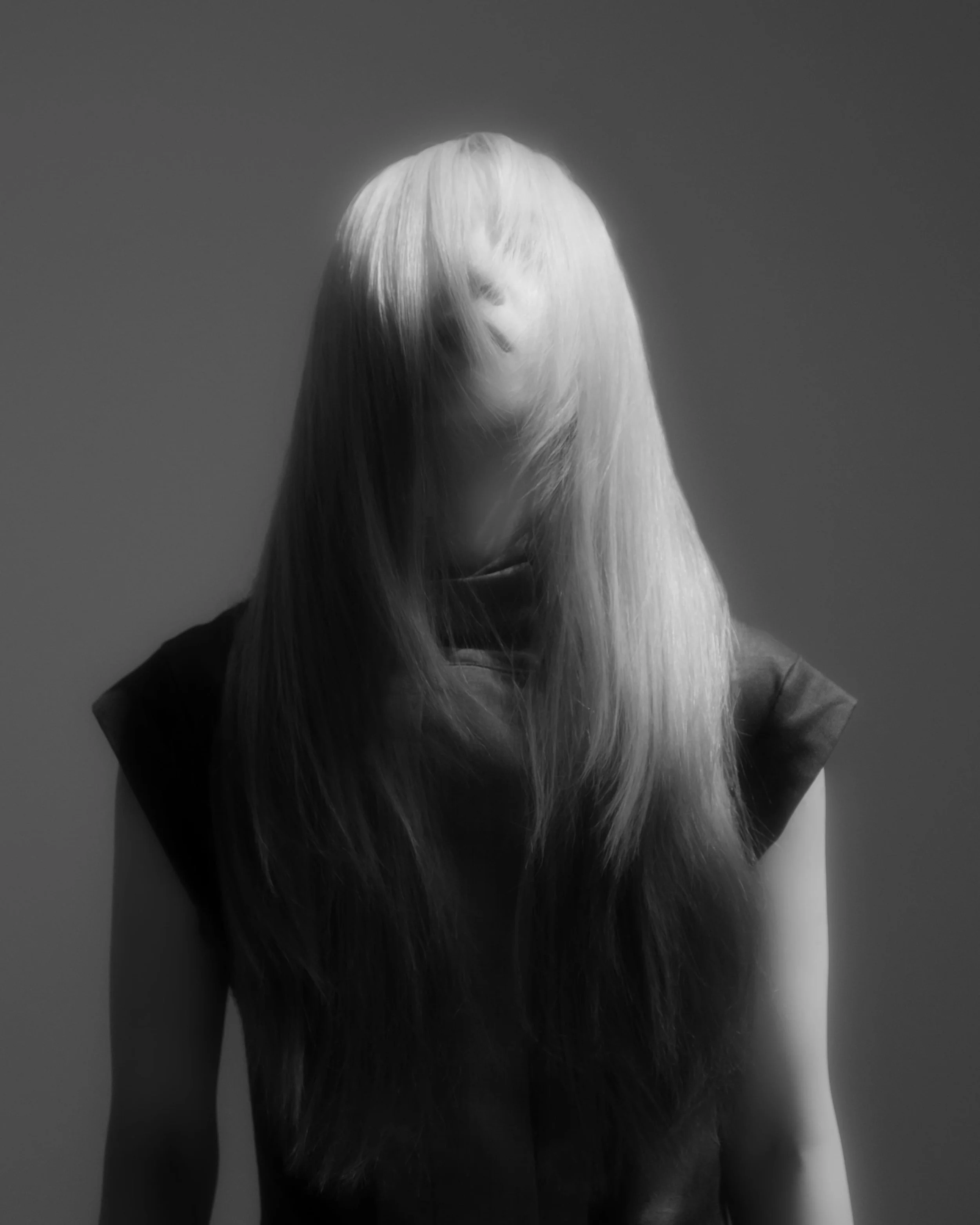A person with long, straight blonde hair covering their face, standing against a plain background, wearing a dark sleeveless top.