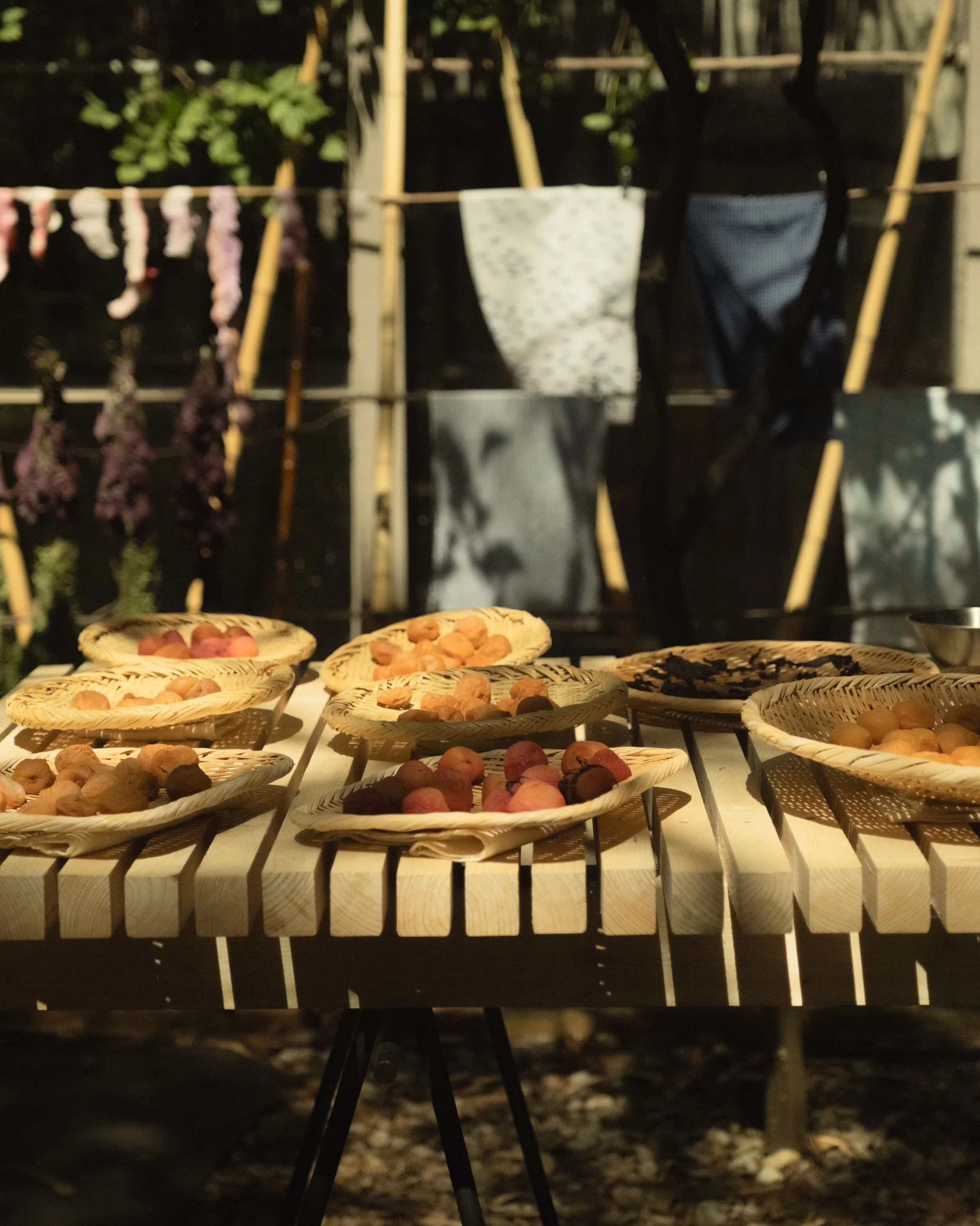 Several woven baskets with small, round fruits on a wooden table outdoors, with laundry hanging on a bamboo clothesline in the background.