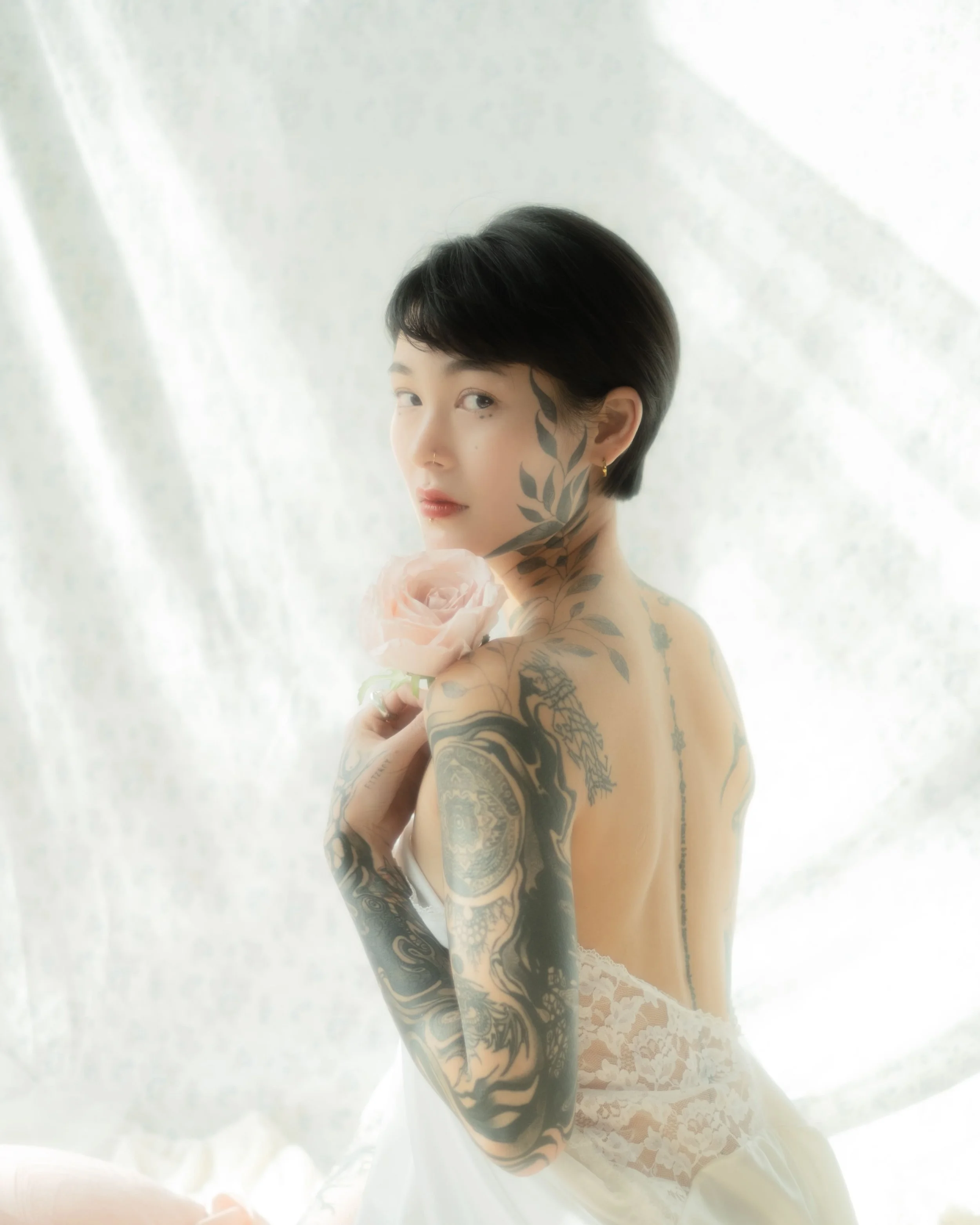 A woman with short black hair, tattoos on her arms and shoulder, and wearing lace underwear, holding a rose, sitting in front of a light-colored background.