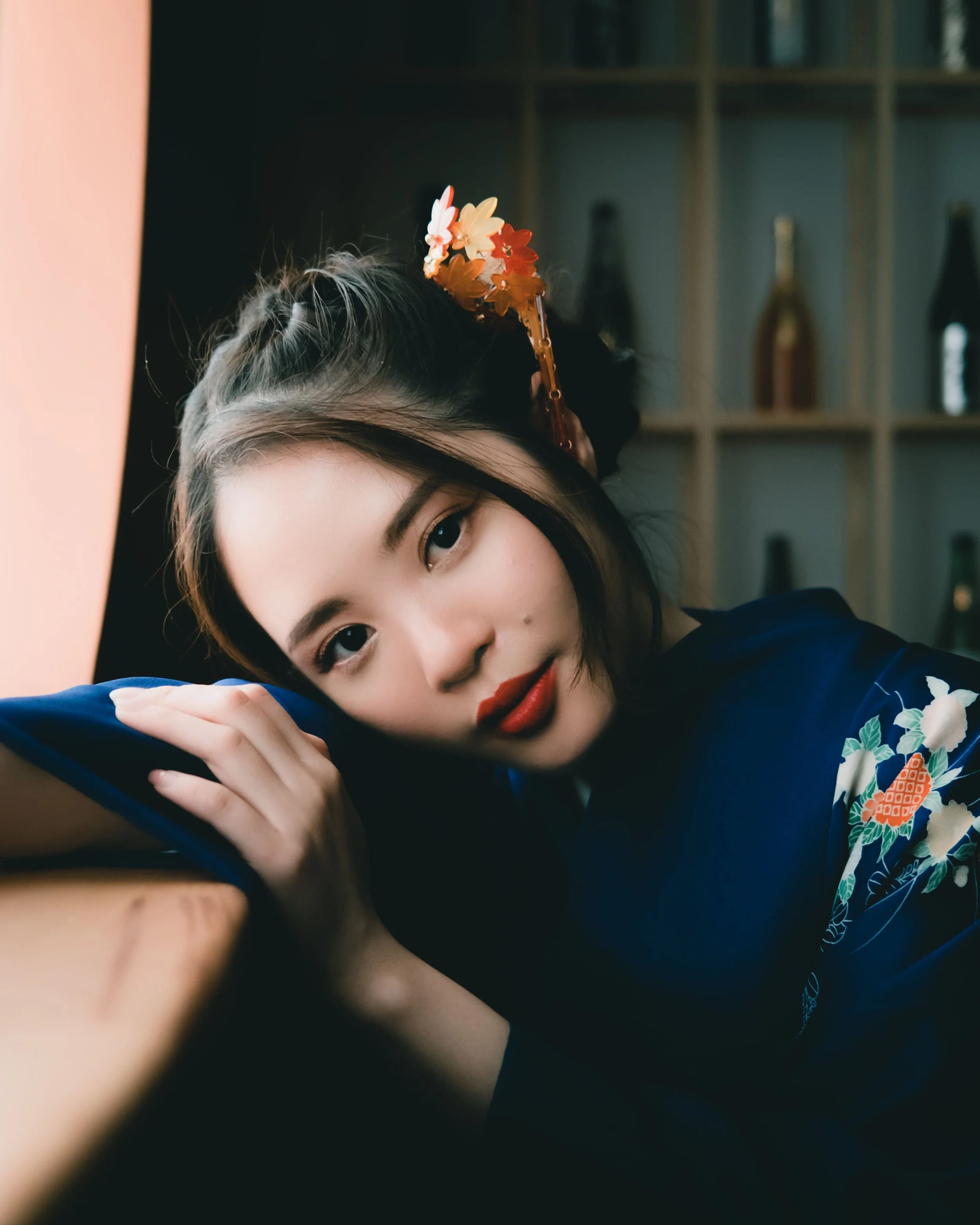 A young woman with dark hair and red lipstick laying on her side, resting her head on her folded arms, with a shelf in the background containing various bottles.