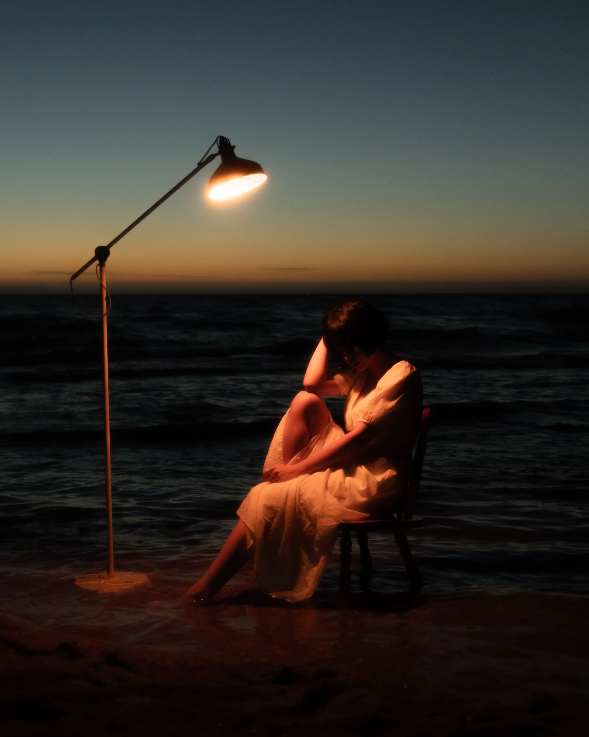 A woman sitting on a chair on the beach at sunset, illuminated by a nearby lamp, with the ocean in the background.