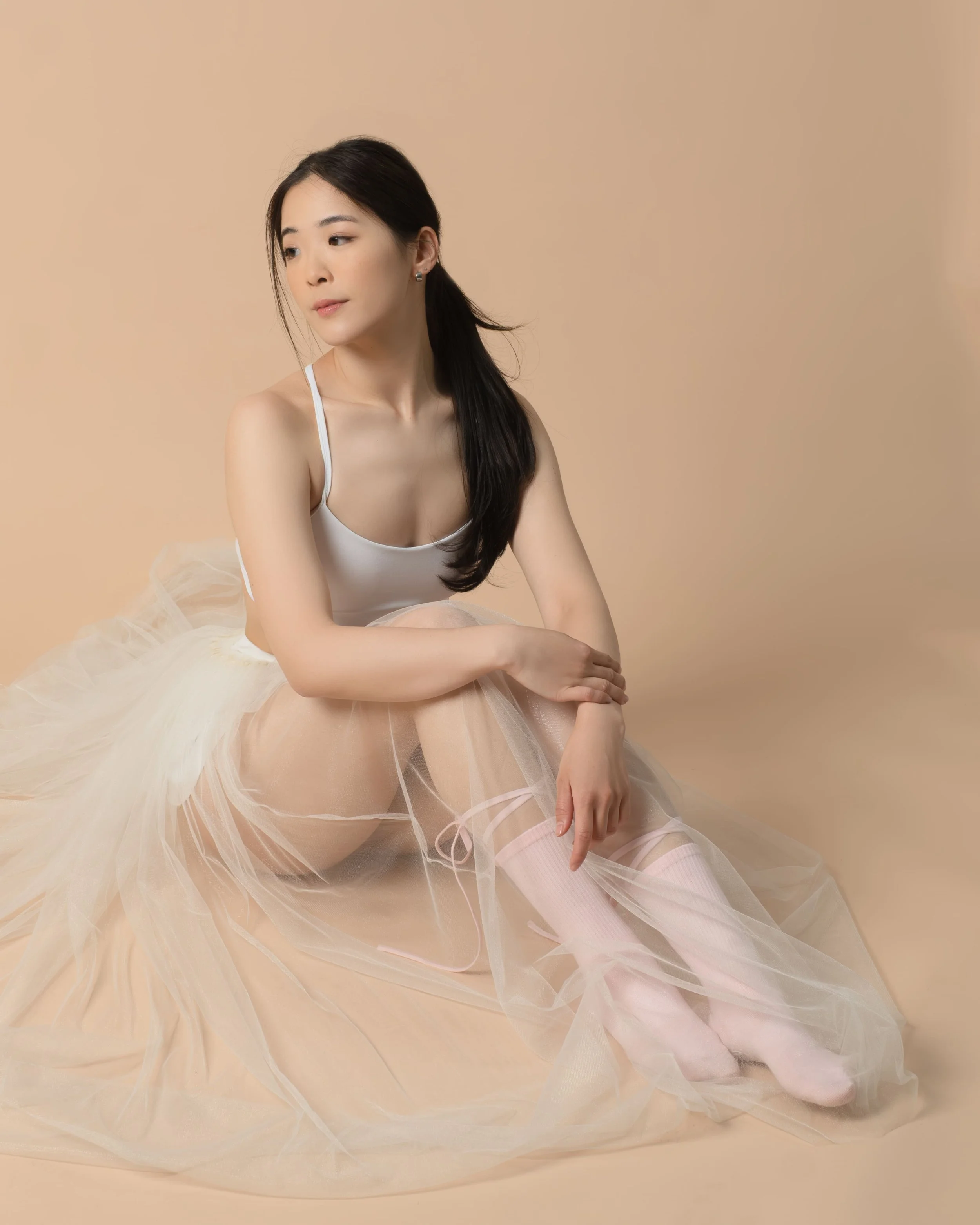 Young woman sitting on the floor, wearing a white sleeveless top, a light-colored tutu skirt, and pink ballet tights with satin ballet shoes, against a beige background.