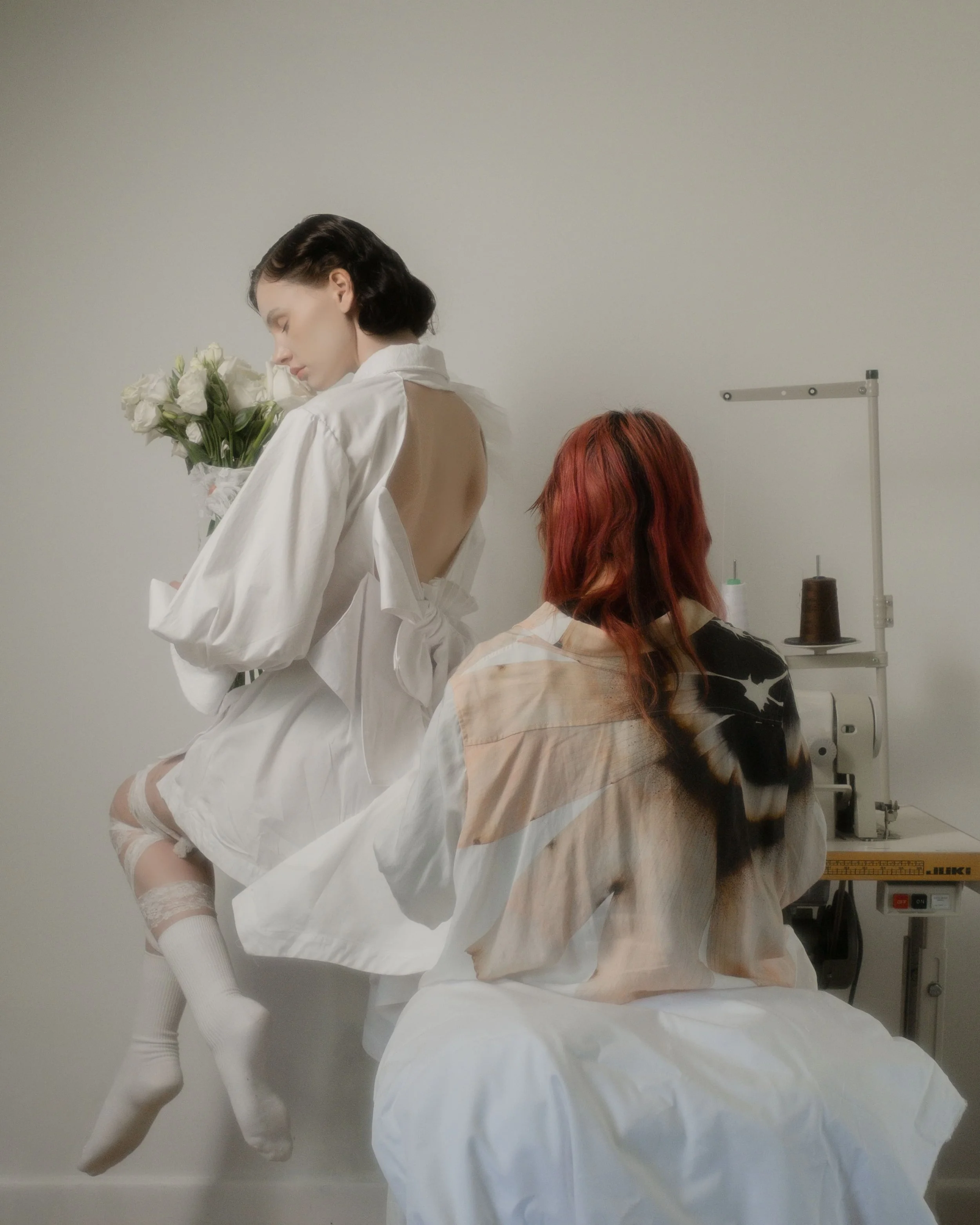Two women in a minimalist room, one sitting on a bed with her back to the camera and the other sitting on the edge holding a bouquet of white roses, with a sewing machine and spools of thread in the background.