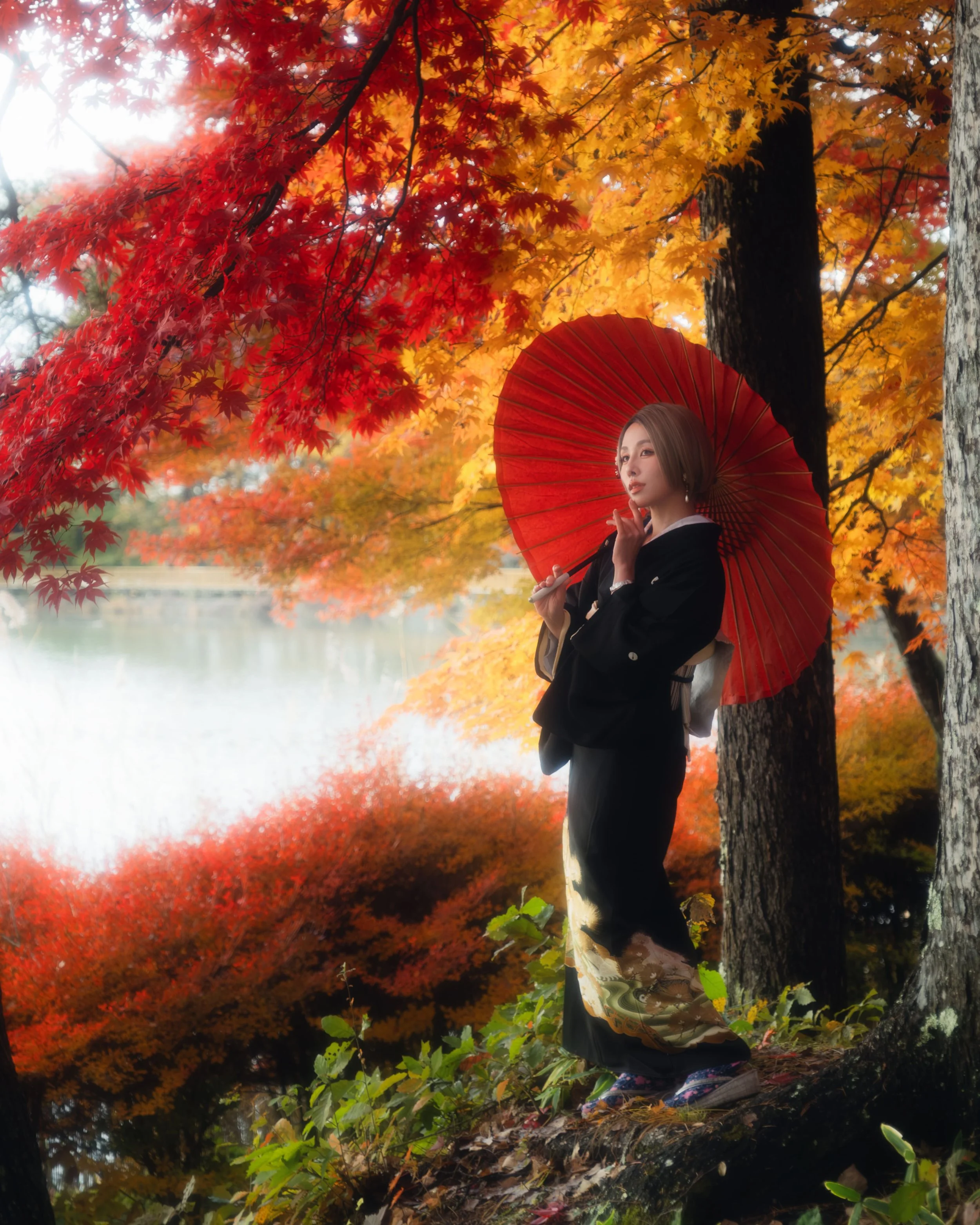 A woman in traditional Japanese attire holding a red parasol, standing among autumn-colored trees with vibrant red, orange, and yellow leaves, near a water body.
