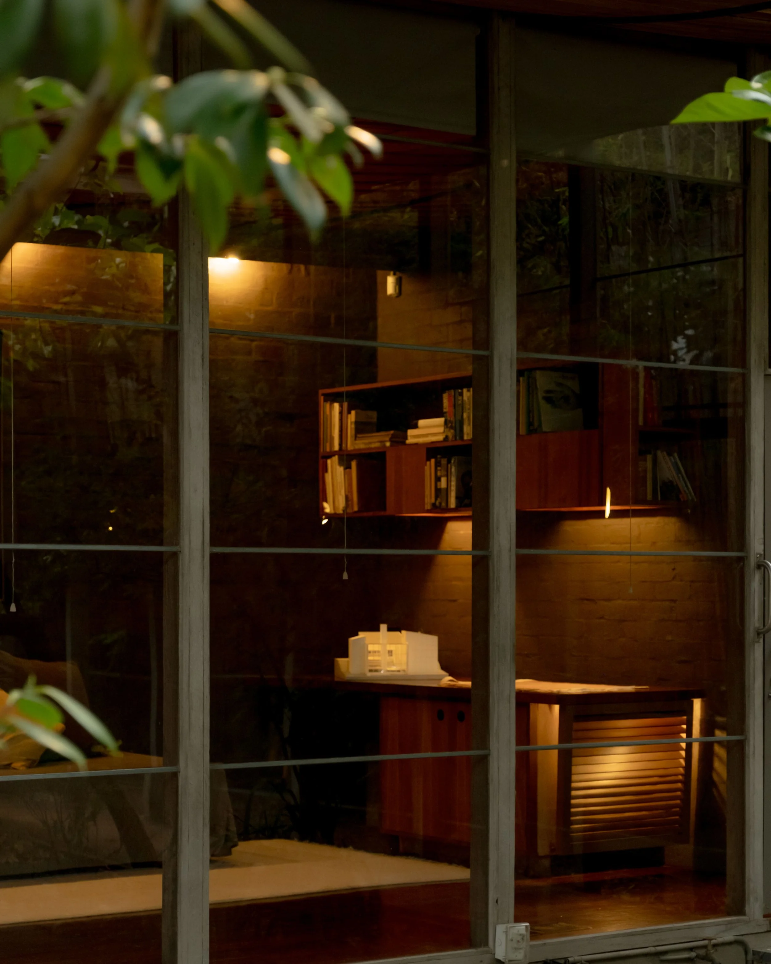 A cozy home office with bookshelves, a desk, and warm lighting, viewed through a glass window with wooden framing, and a few green leaves in the foreground.