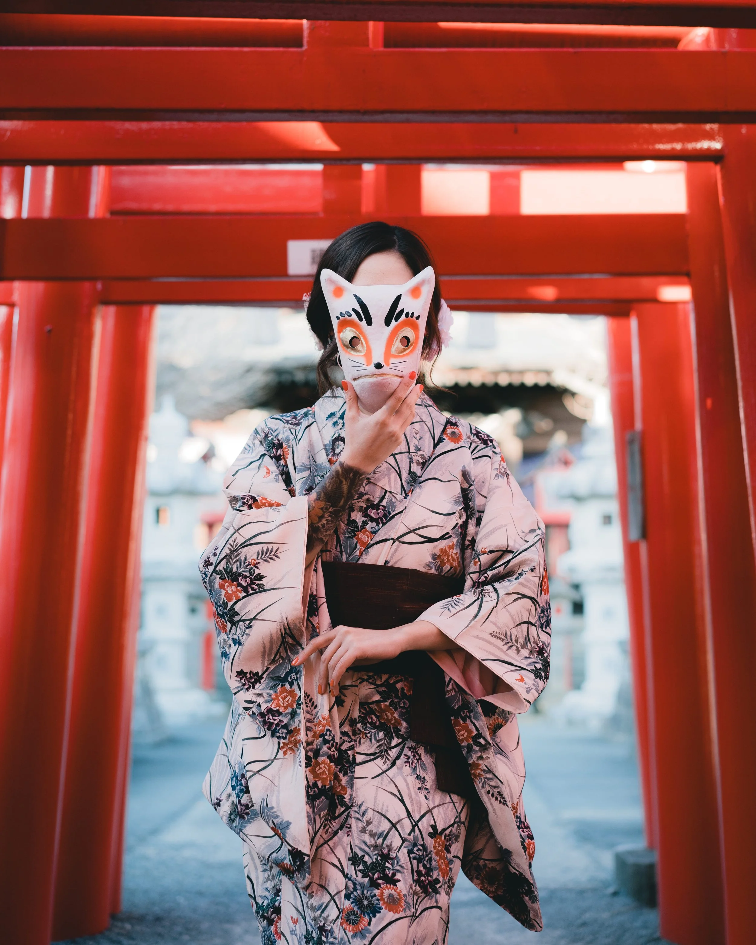 A woman in a floral kimono holds a fox mask in front of her face in front of red paper torii gates.