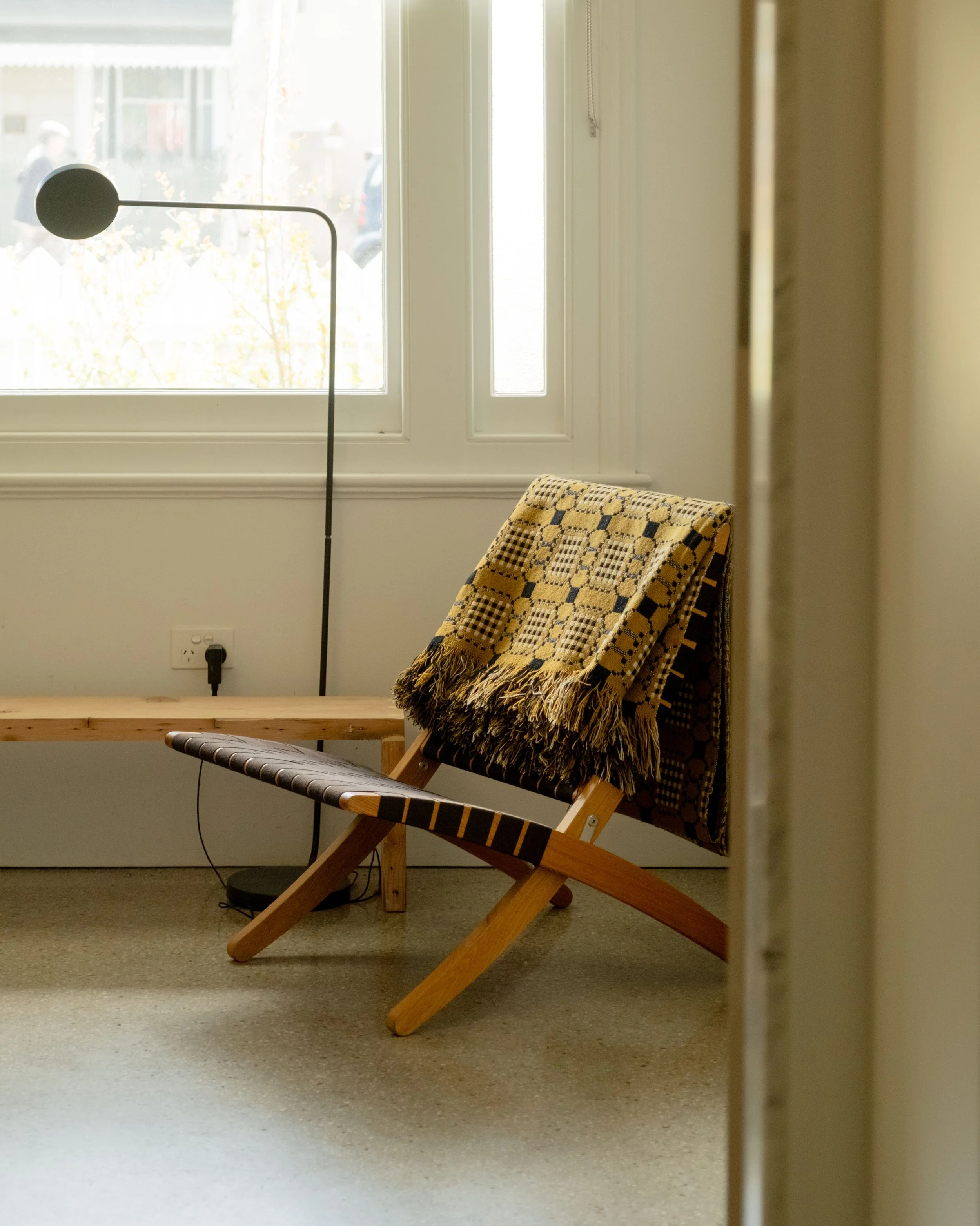 A cozy wooden lounge chair with a patterned blanket draped over it, positioned next to a window with sunlight shining through, in a minimalist room.
