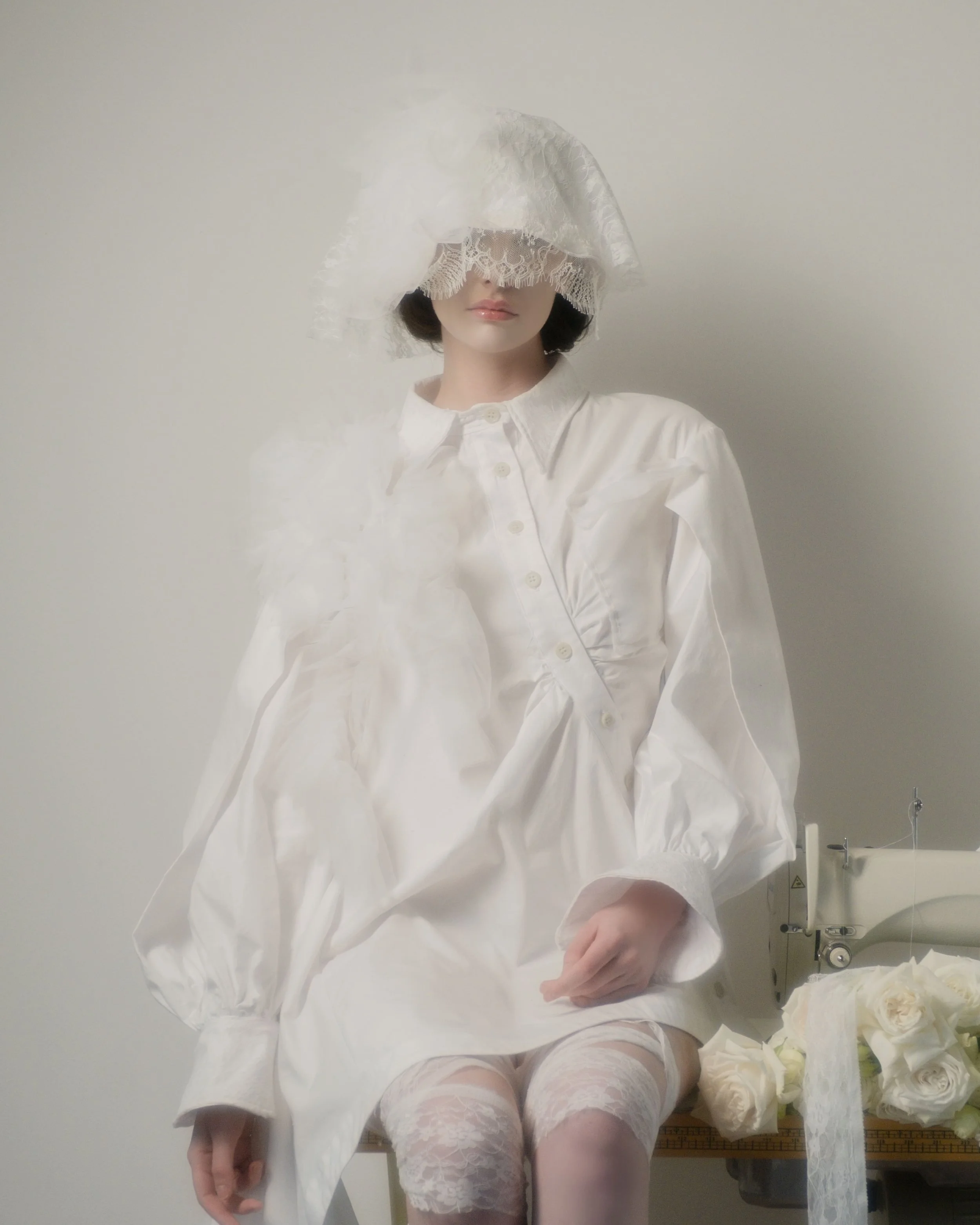 A woman dressed in a white blouse with lace and lace tights, sitting on a table next to a sewing machine surrounded by white roses, with a lacy bonnet covering part of her face.