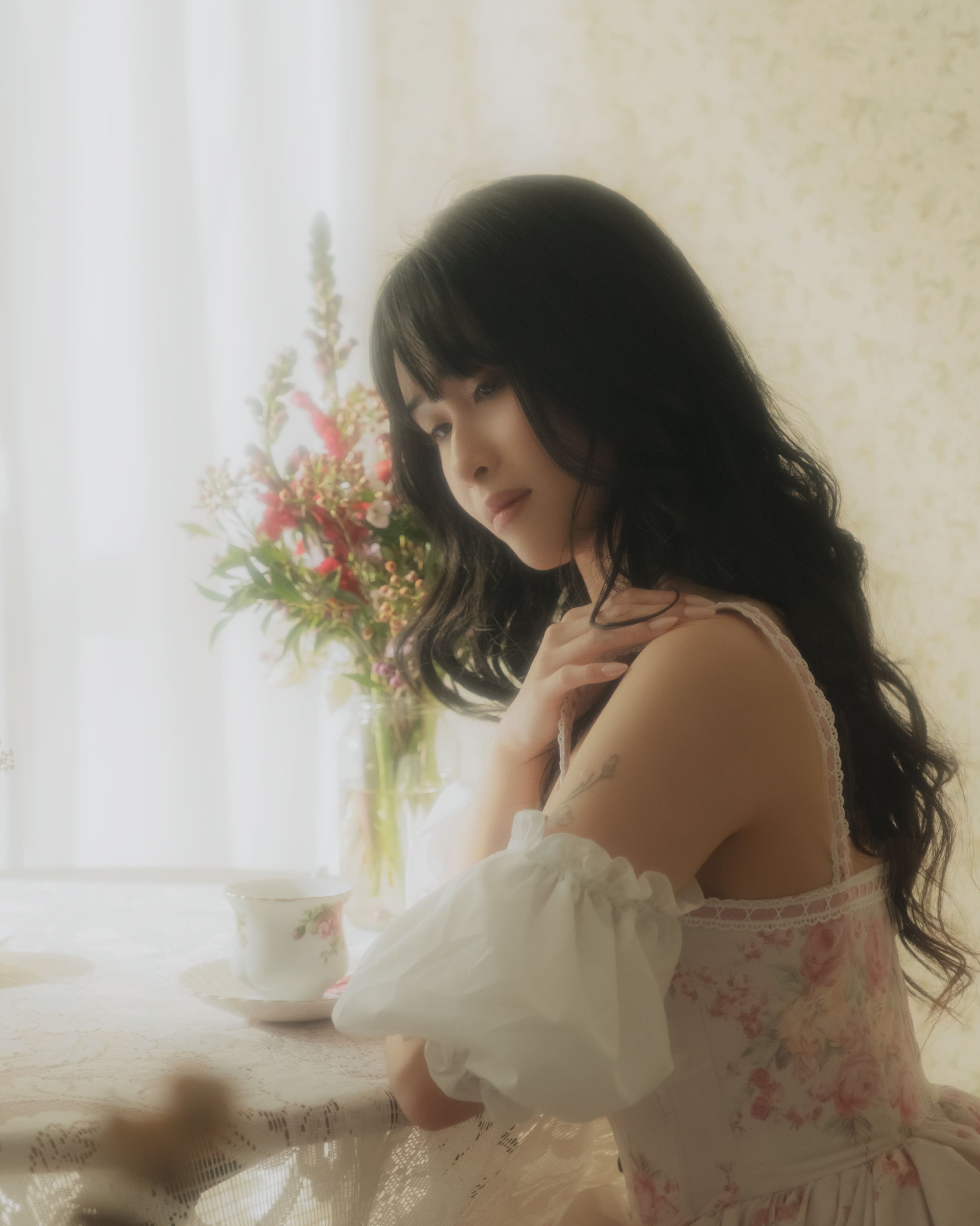 A woman with black hair and bangs gazes downward, touching her shoulder, with a background of a floral arrangement and lace curtains.