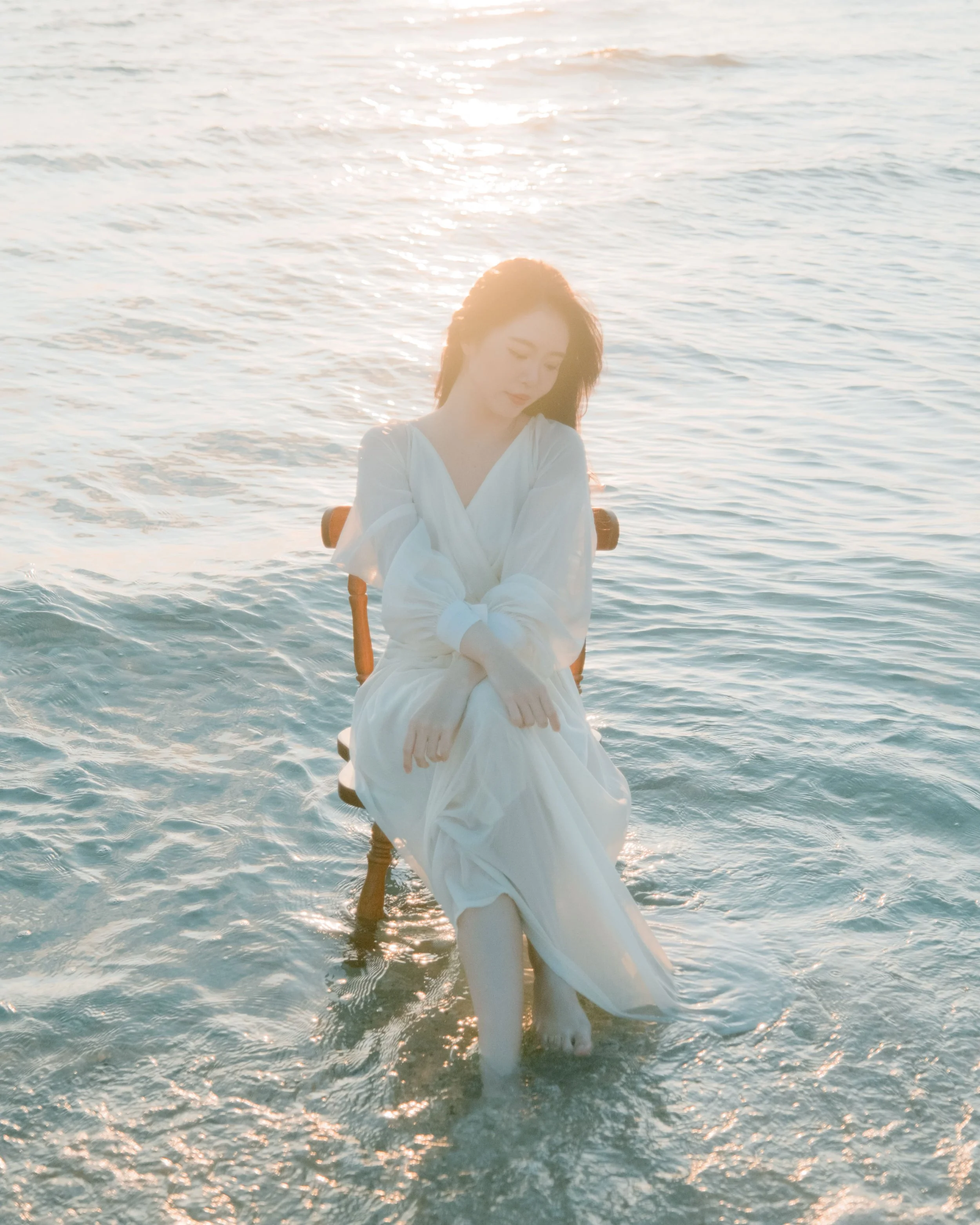 A woman in a white dress sitting on a chair in shallow ocean water during sunset, with her eyes closed and her hands resting on her knees.