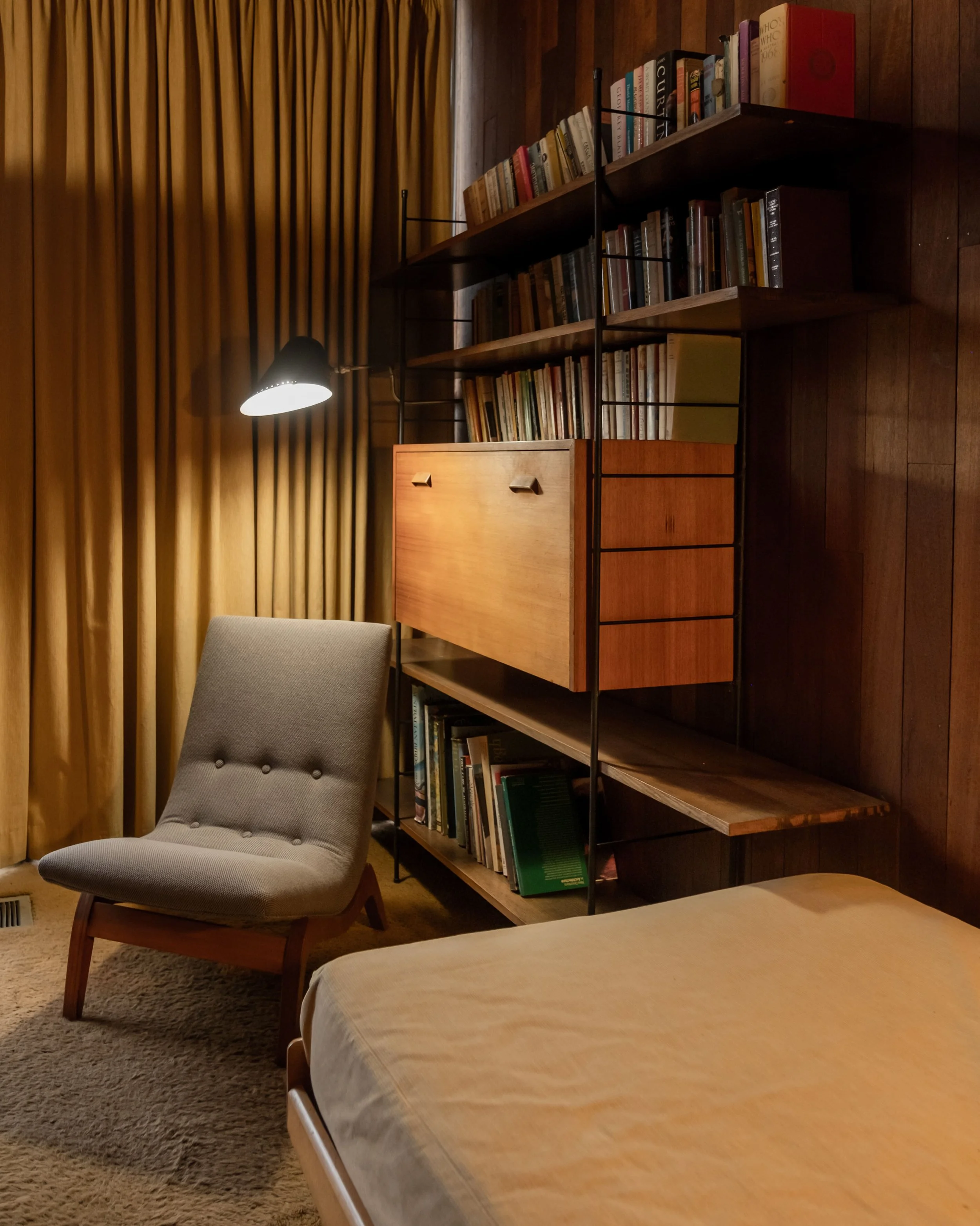 A cozy room with a bed, a gray upholstered lounge chair, a wooden bookshelf with books, a black wall-mounted reading lamp, and gold curtains.