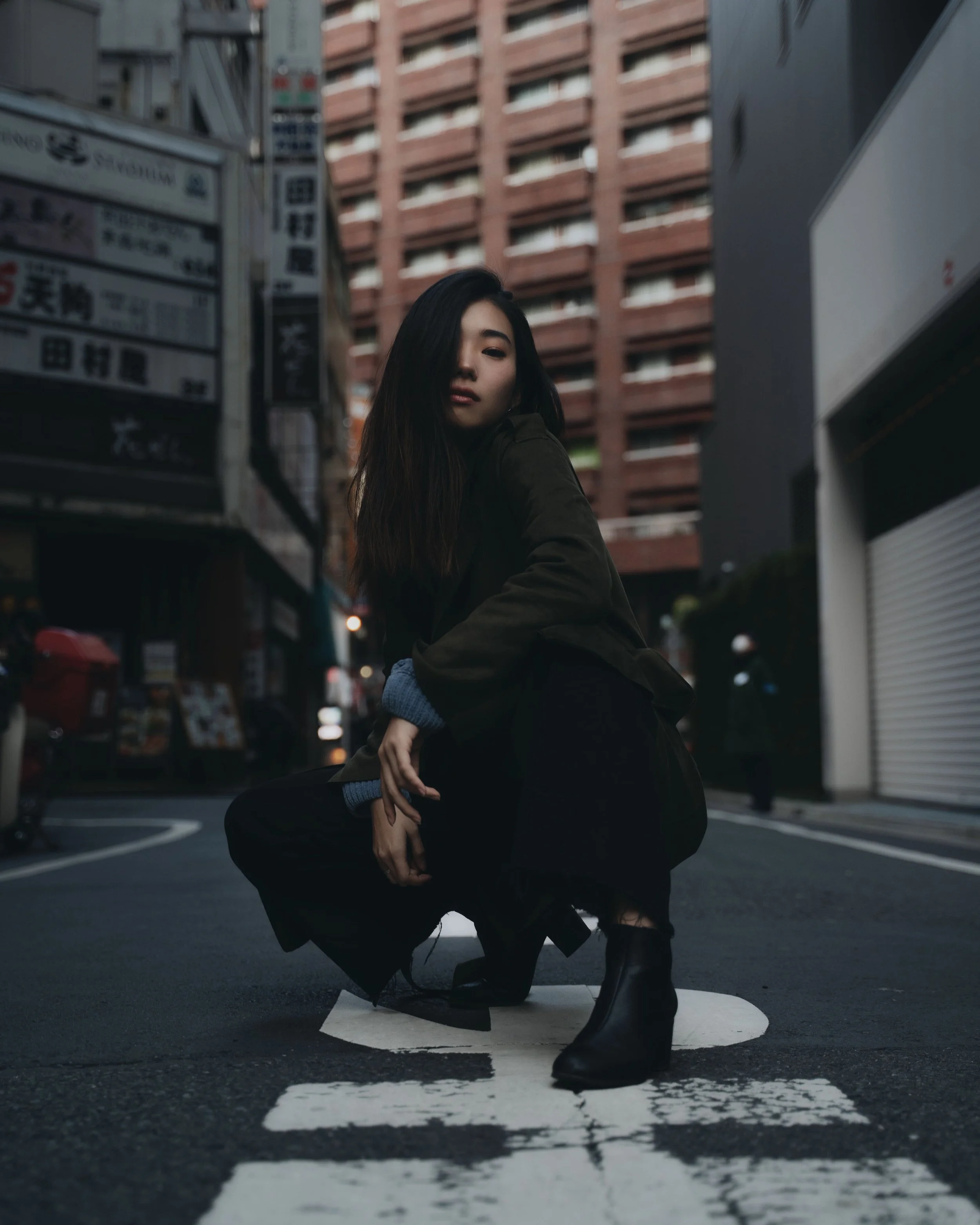 A woman in black boots crouching on a city street at dusk, with tall buildings and storefront signs in the background.