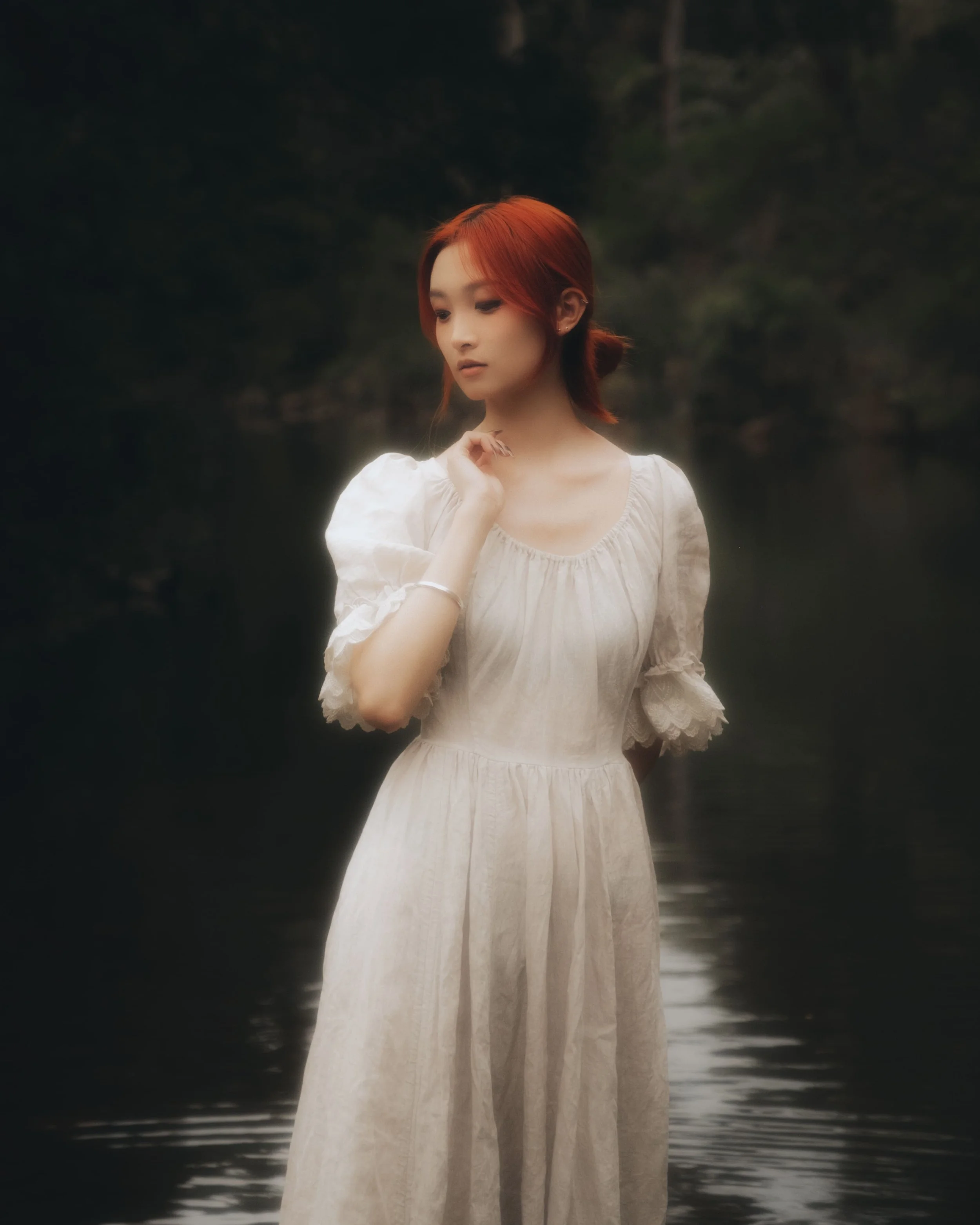 A young woman with red hair in a white vintage-style dress standing near a body of water with dark, blurred trees in the background.