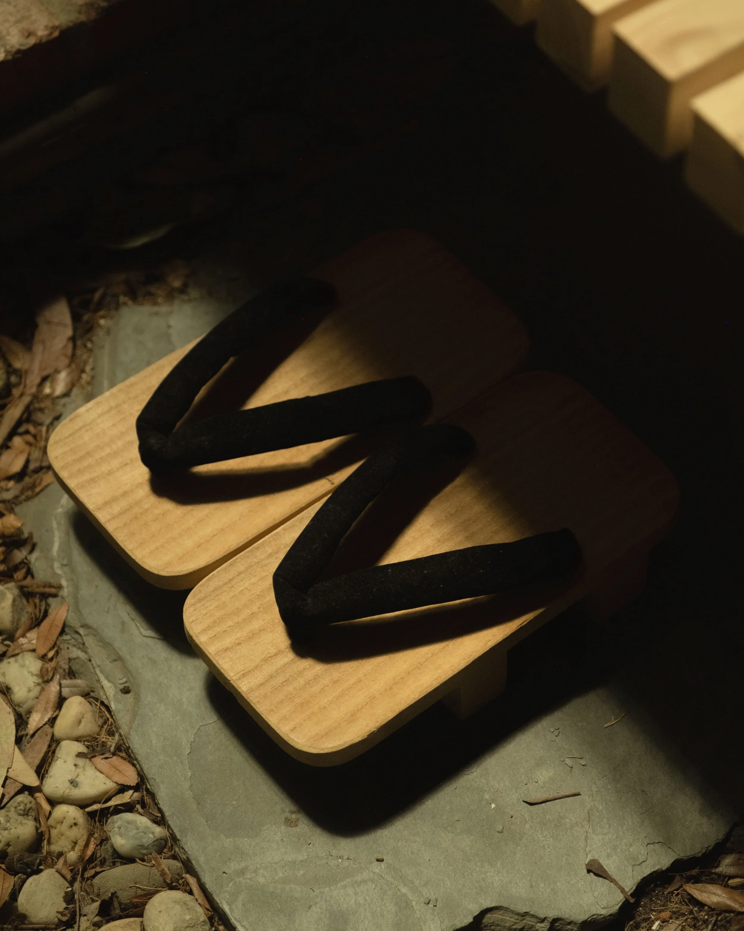 Pair of wooden slide sandals, illuminated by sunlight, resting on a concrete surface with small rocks and dry leaves around.