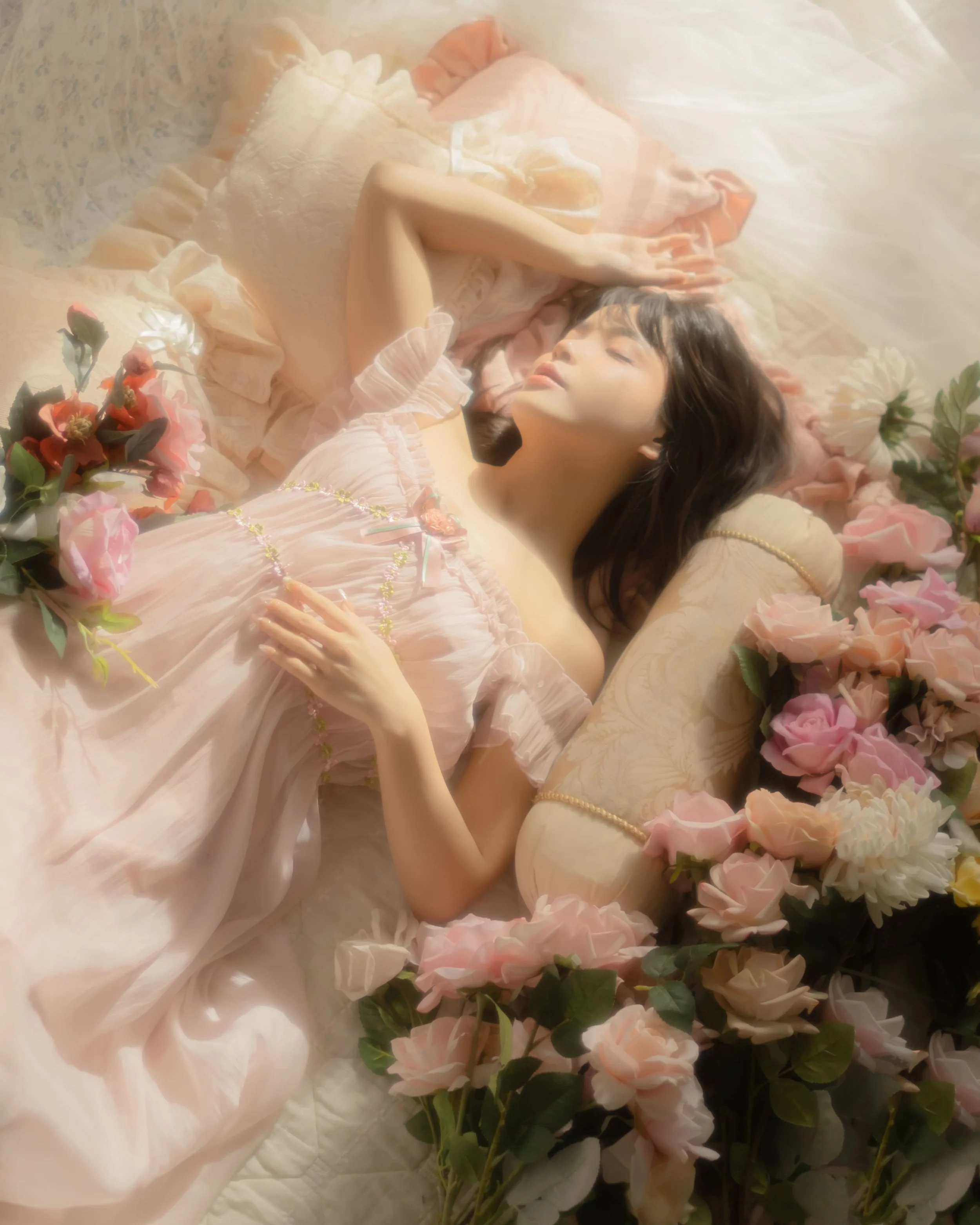 A woman lying on a bed surrounded by pink and peach roses, wearing a pink dress, with her eyes closed and one arm resting on her forehead.