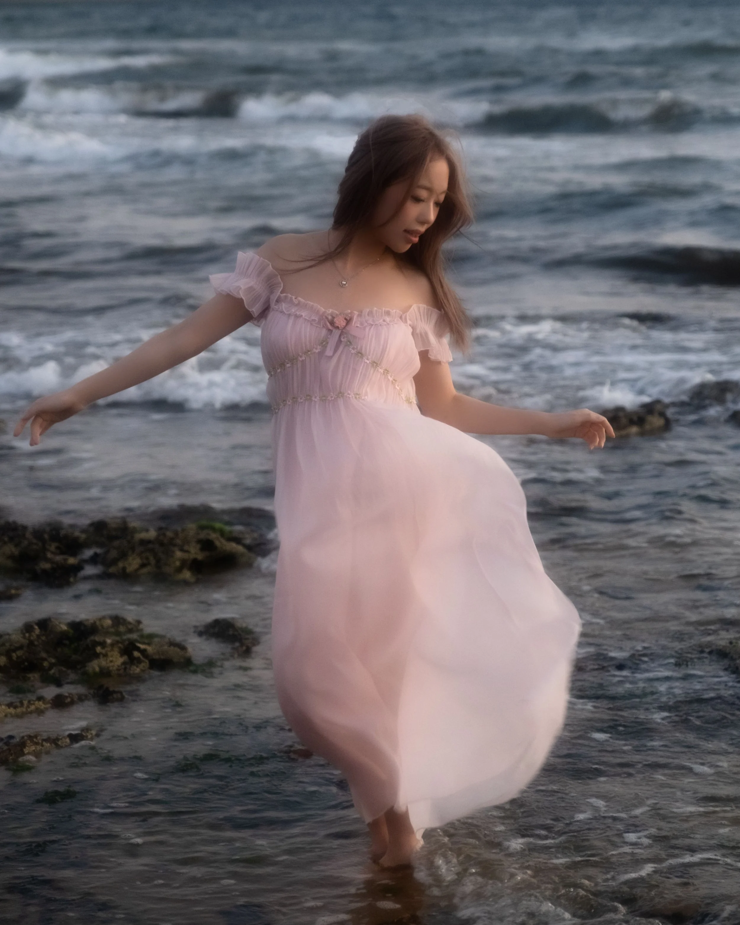A woman in a light pink, off-shoulder dress with ruffles and a small bow, walking in the ocean with water reaching her ankles, as the waves gently roll in the background.