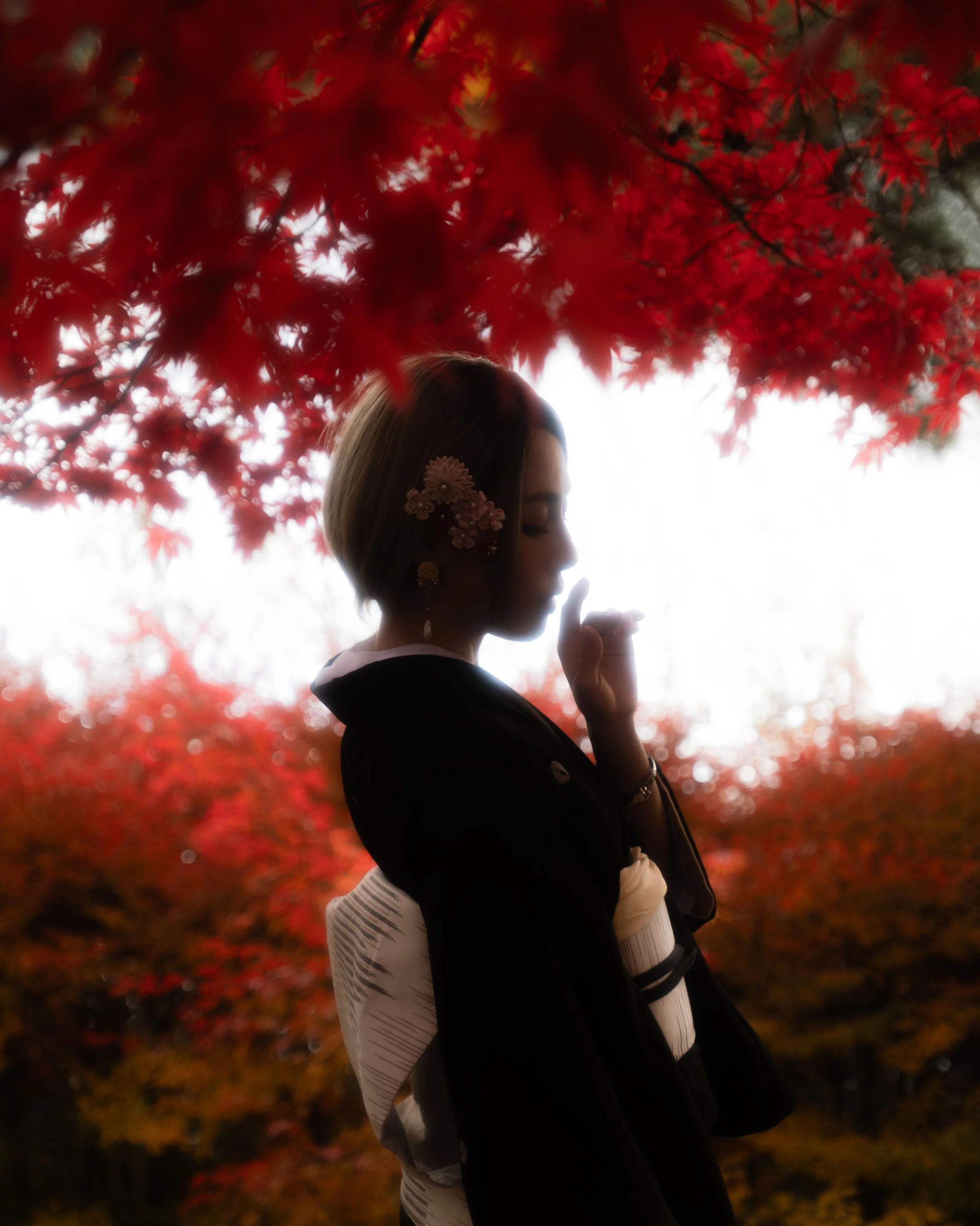 A woman with floral hair accessories and earrings holds a vape in her right hand. She is dressed in black and white clothing and is positioned in front of vibrant red autumn leaves, with her face in profile against a bright sky background.