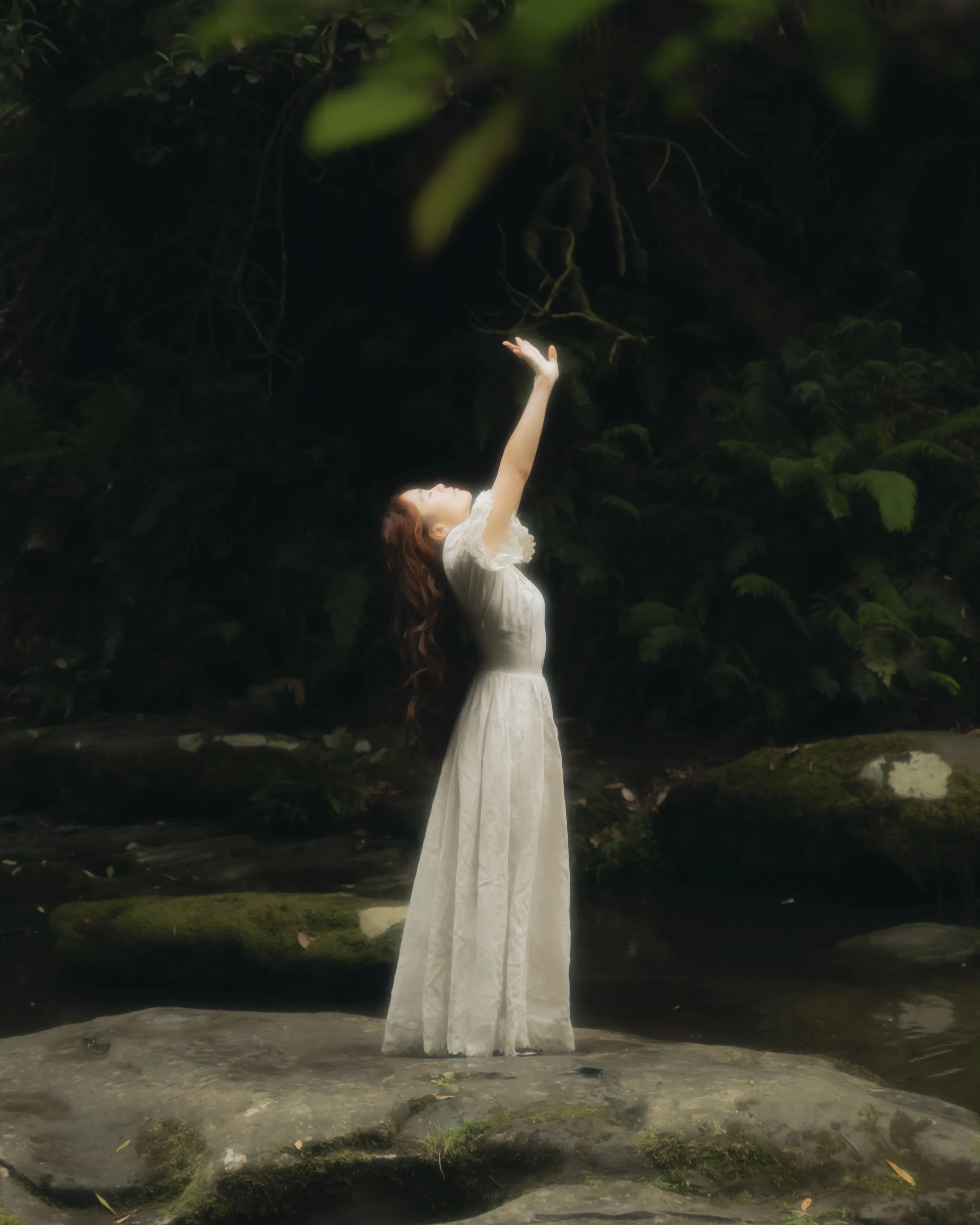 Woman in a white dress with long wavy hair standing on a rock by a creek, reaching upward with her arms in front of a dark, leafy background.