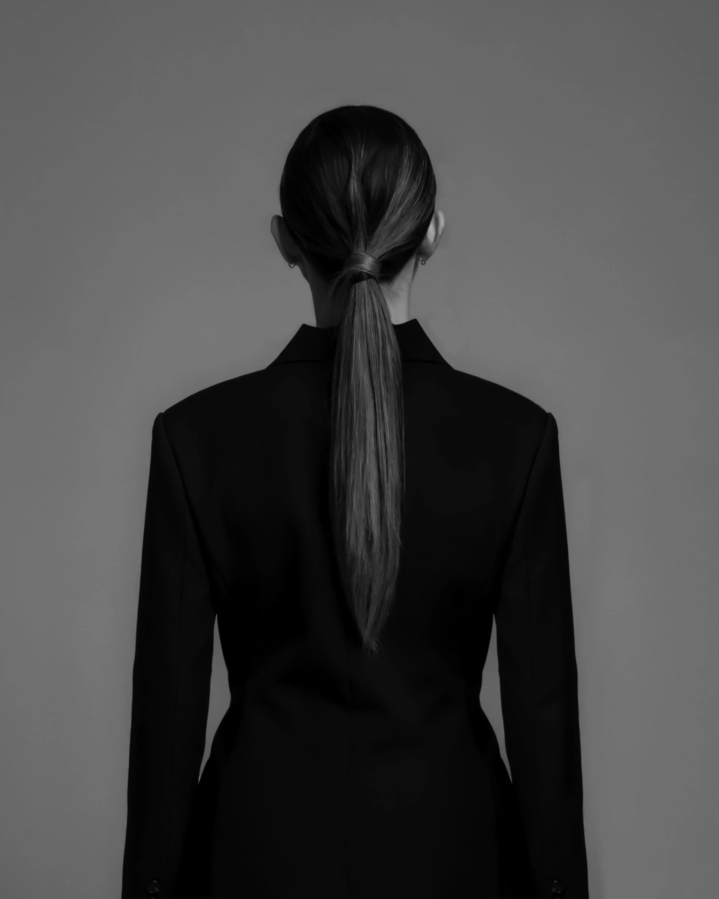 Back view of a woman with long hair tied in a ponytail, wearing a formal black suit during a photoshoot.