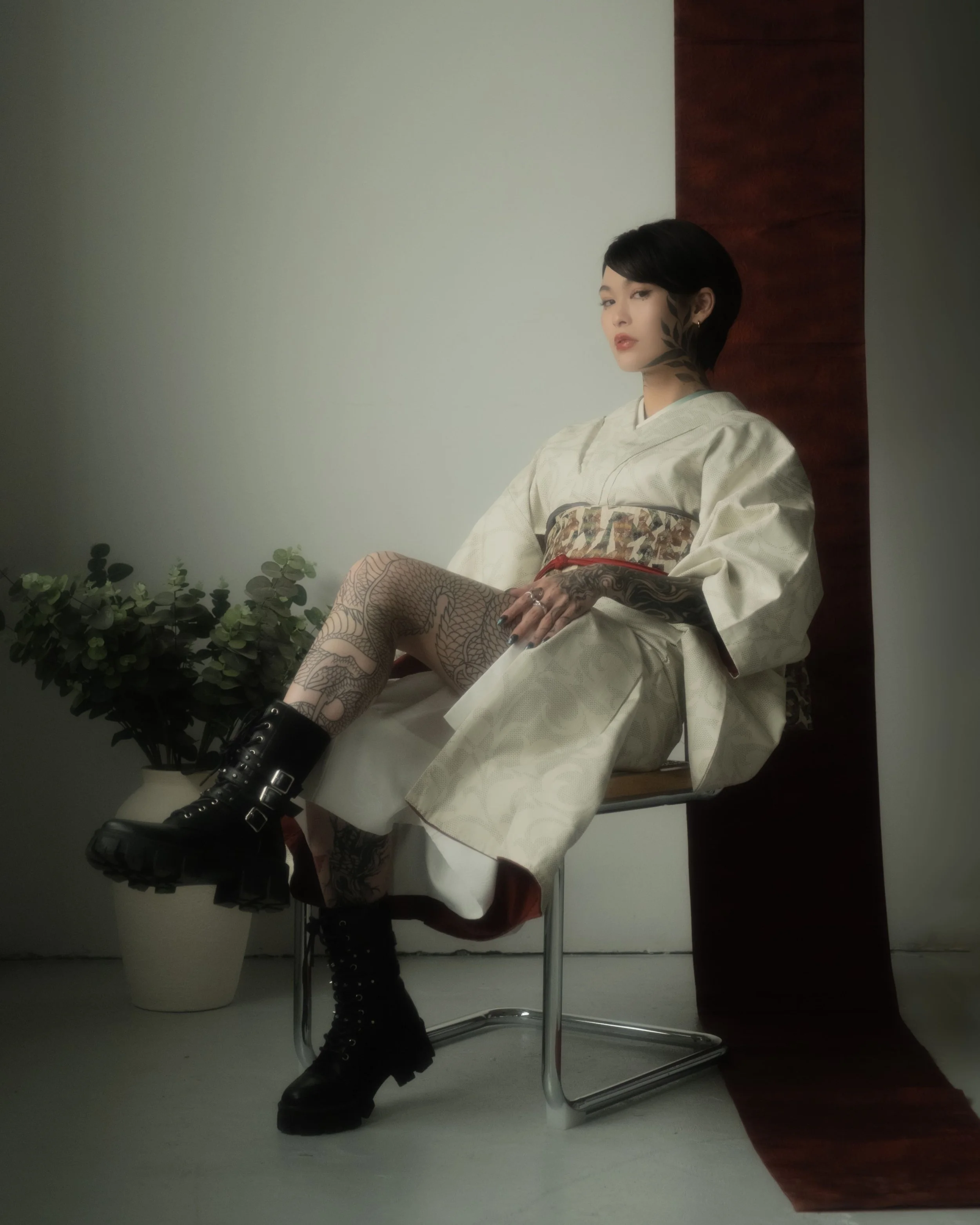 A woman with short black hair and tattoos, wearing combat boots and a traditional kimono, sitting on a modern wooden chair, with a plant and curtain in the background.