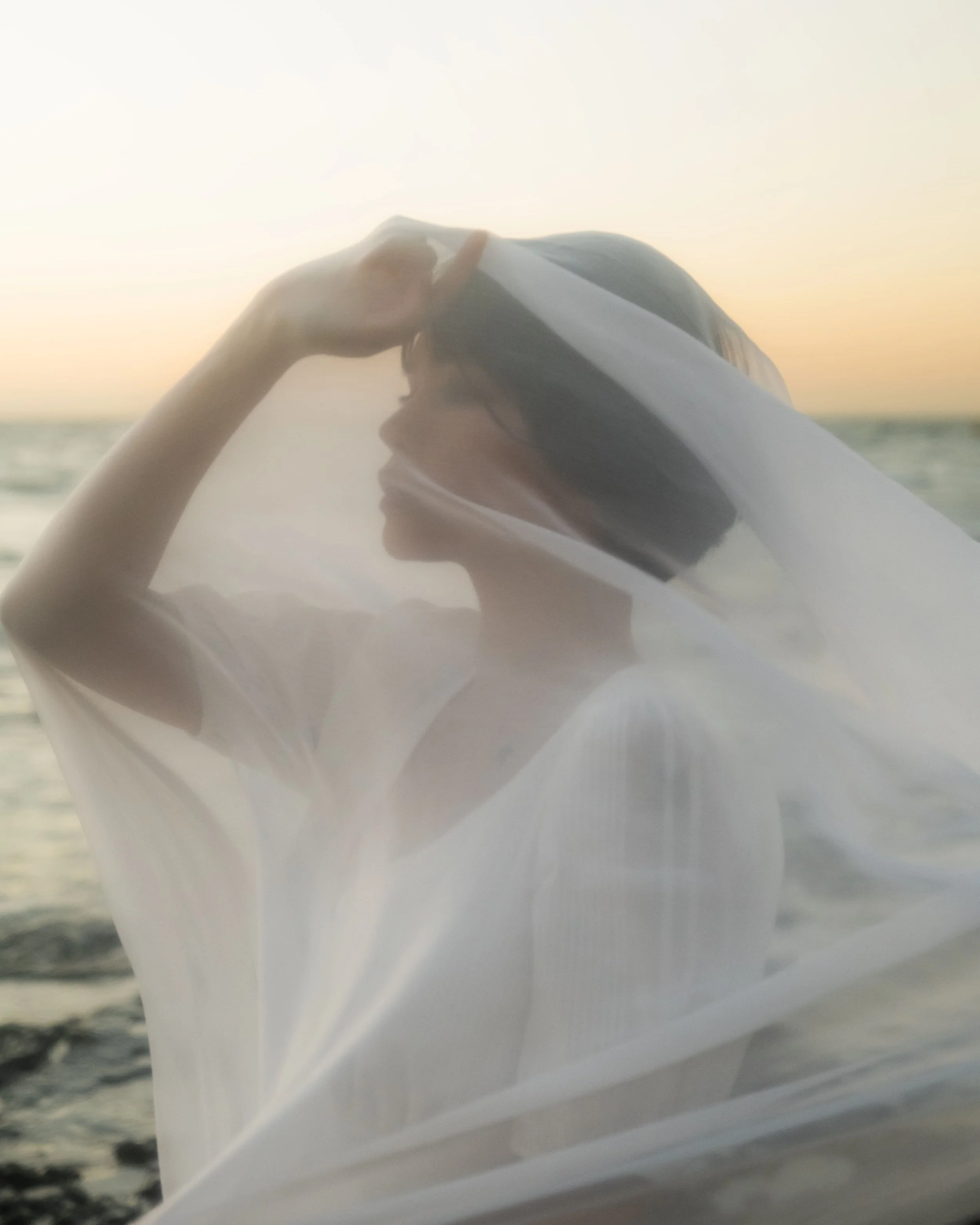 Woman with short black hair standing near the ocean at sunset, covered partially by a sheer white fabric.