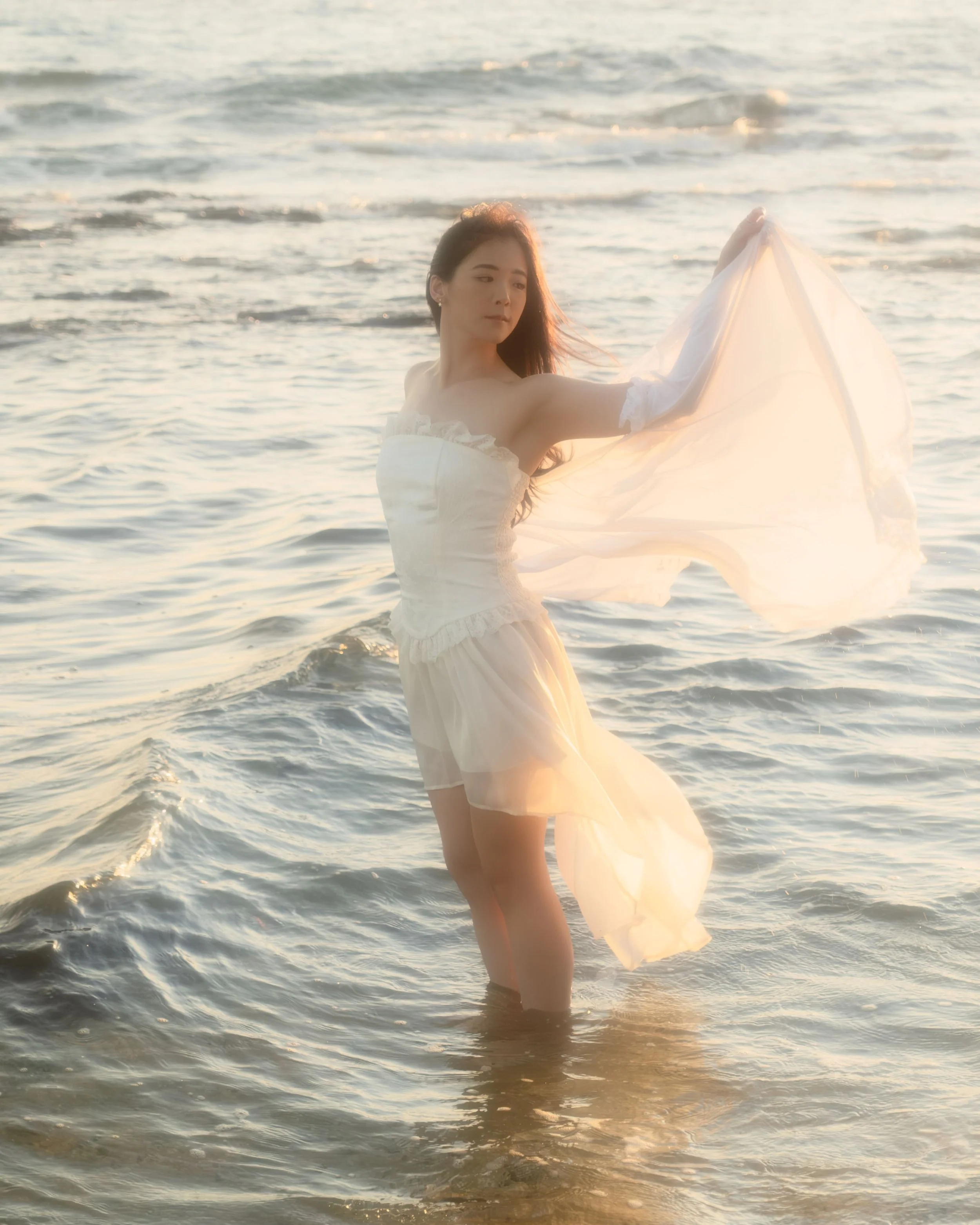 Woman in a white dress standing in the ocean at sunset, holding a flowing piece of fabric, with water reaching her knees.