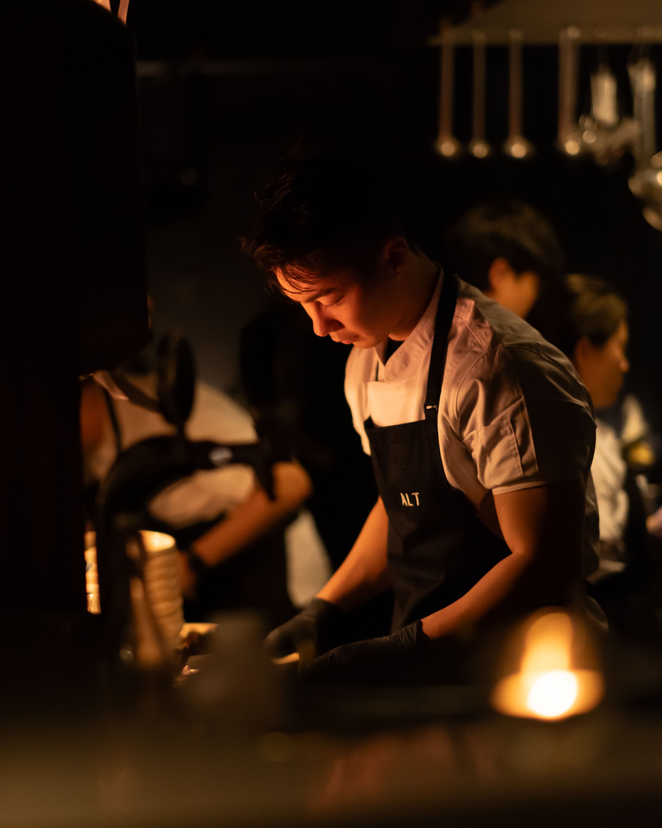 A young chef wearing a black apron with the letter 'ALT' working in a dimly lit kitchen, preparing food with others in the background.
