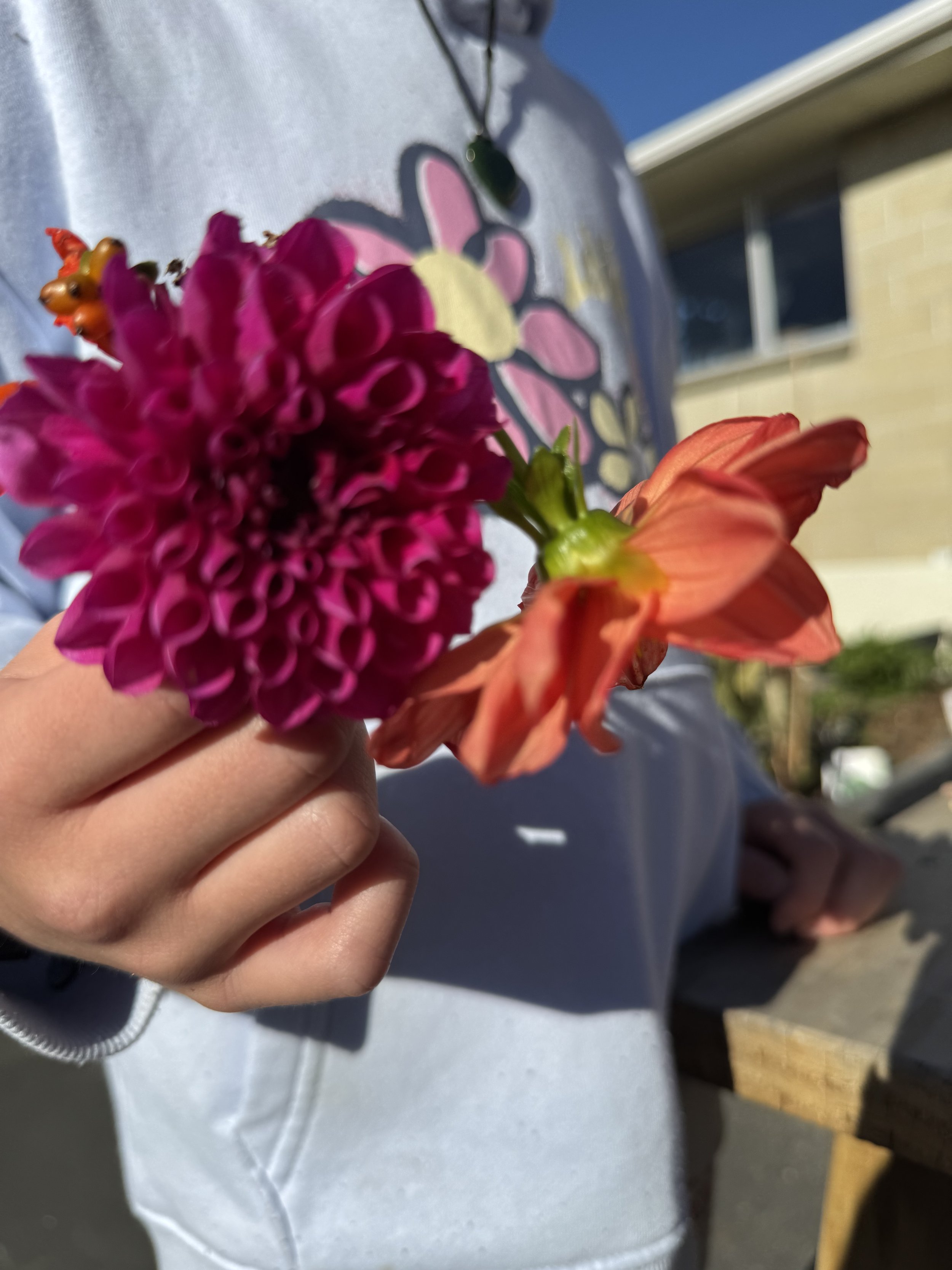 Person holding a pink and orange flower bouquet, wearing a white shirt with flower design, standing outdoors.