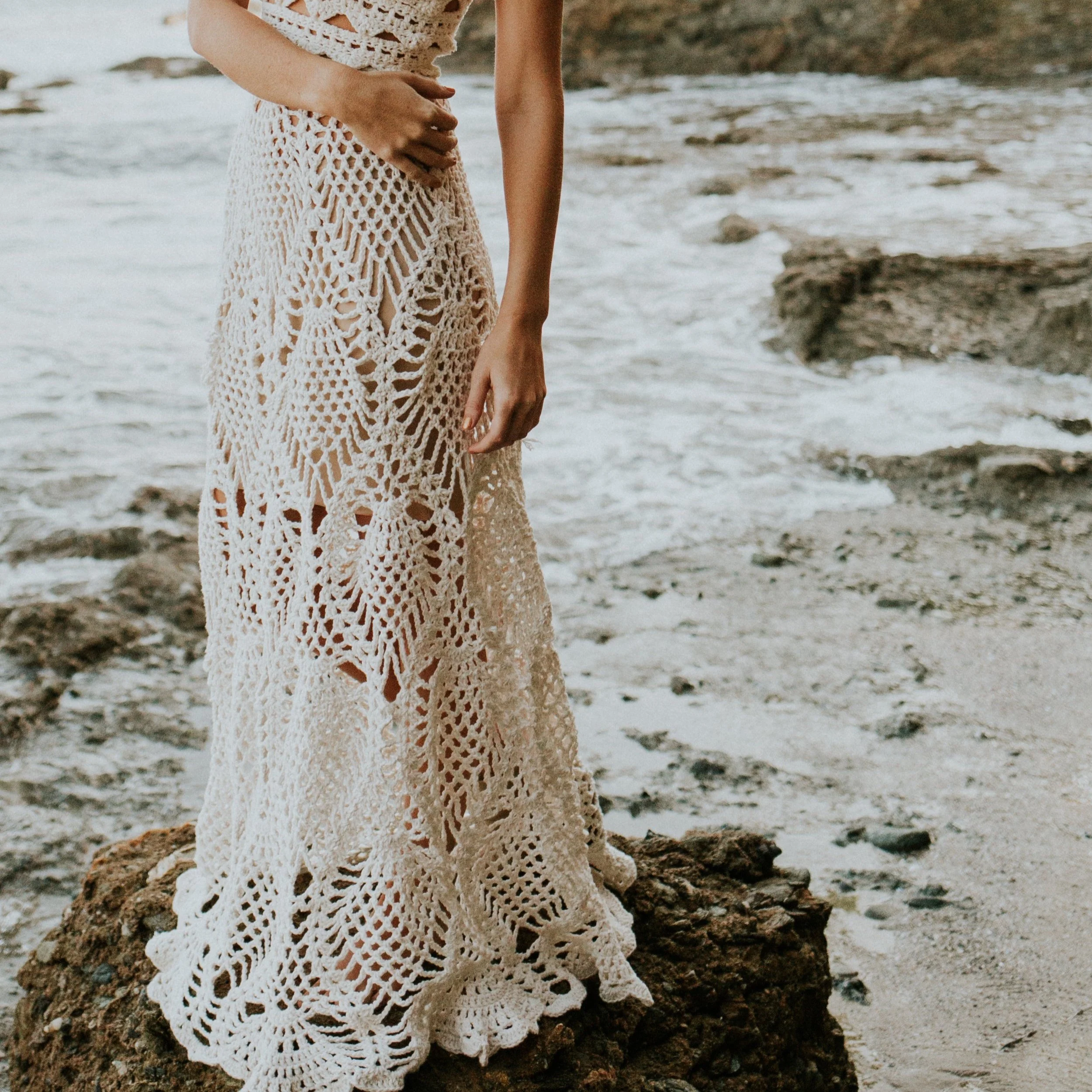 Close-up of a woman wearing a crocheted dress standing on a rock by the ocean shoreline.