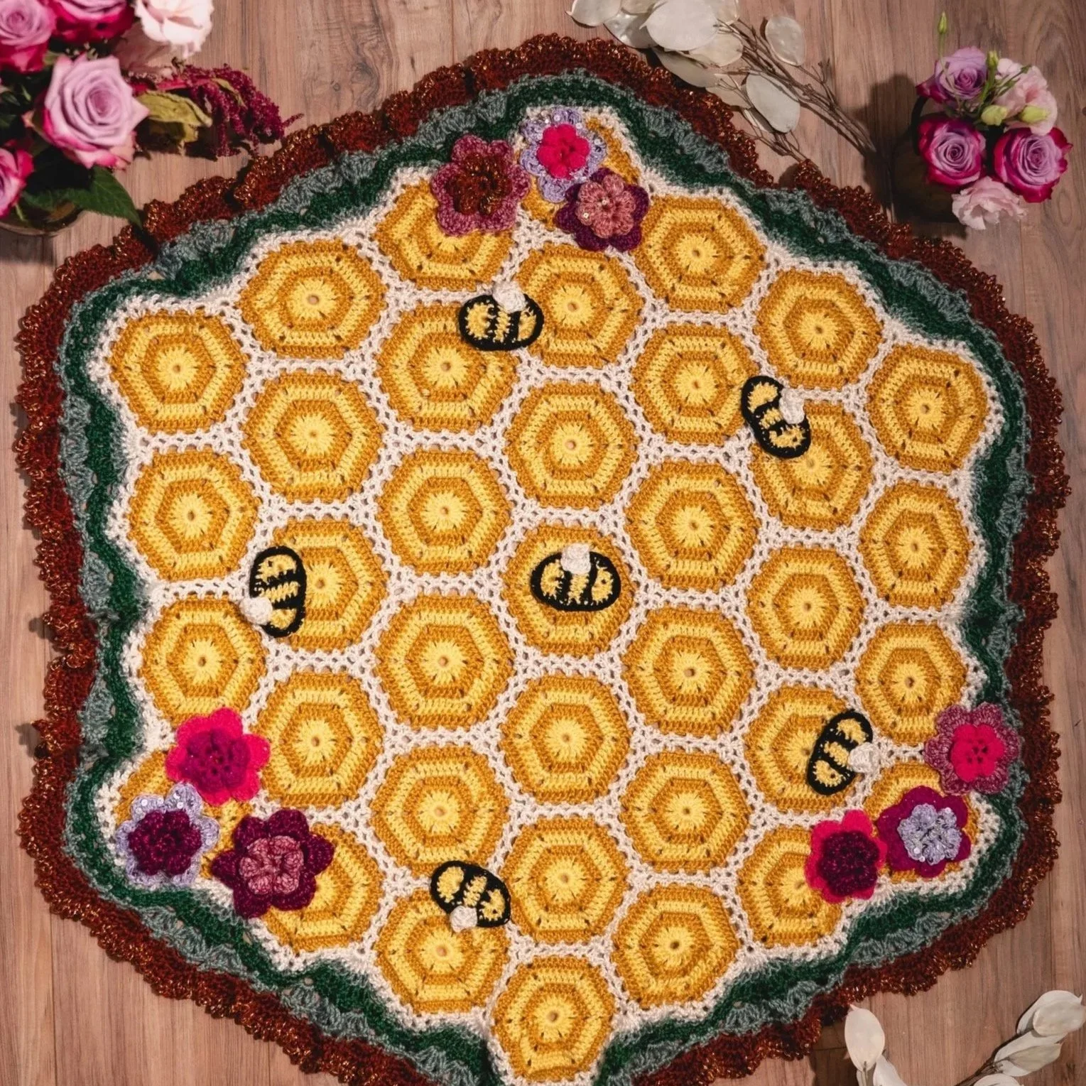 A circular, decorative crochet rug featuring a honeycomb pattern with bees and floral accents, placed on a wooden floor, with surrounding flowers.