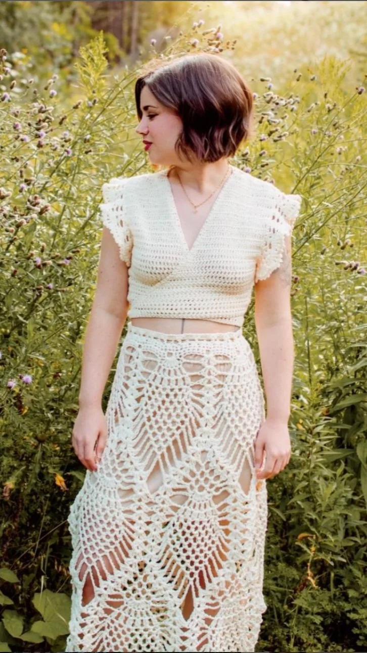 Woman in cream crochet cap sleeve crop top and matching maxi skirt with intricate lace patterns, standing in a sunny meadow