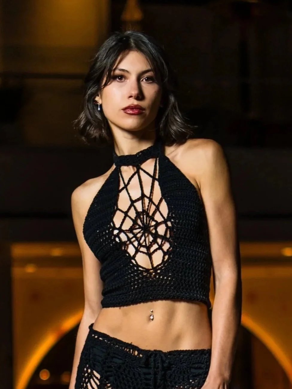 Young woman with shoulder-length dark hair wearing a black crocheted halter top with a spiderweb design in the front, standing indoors with warm background lighting.