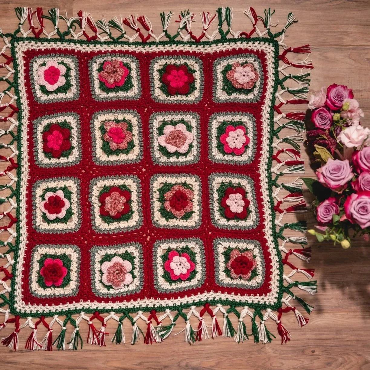 A colorful crochet tablecloth with a floral pattern and fringed edges, placed on a wooden floor next to a bouquet of pink and white roses.
