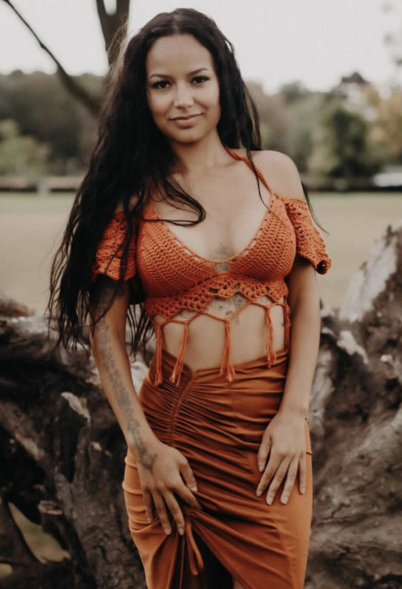 A woman with long dark hair standing outdoors in front of a tree and rocks, wearing an orange crochet top and a matching skirt.