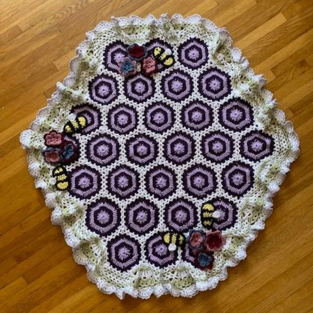 Round crocheted blanket with purple and white hexagon pattern, decorated with crocheted bees and flowers, on a wooden floor.