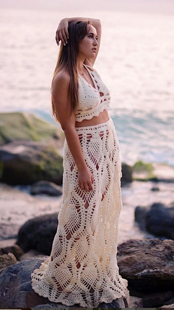 Woman in cream crochet halter crop top and matching maxi skirt posing on seaside rocks during sunset with ocean in the background