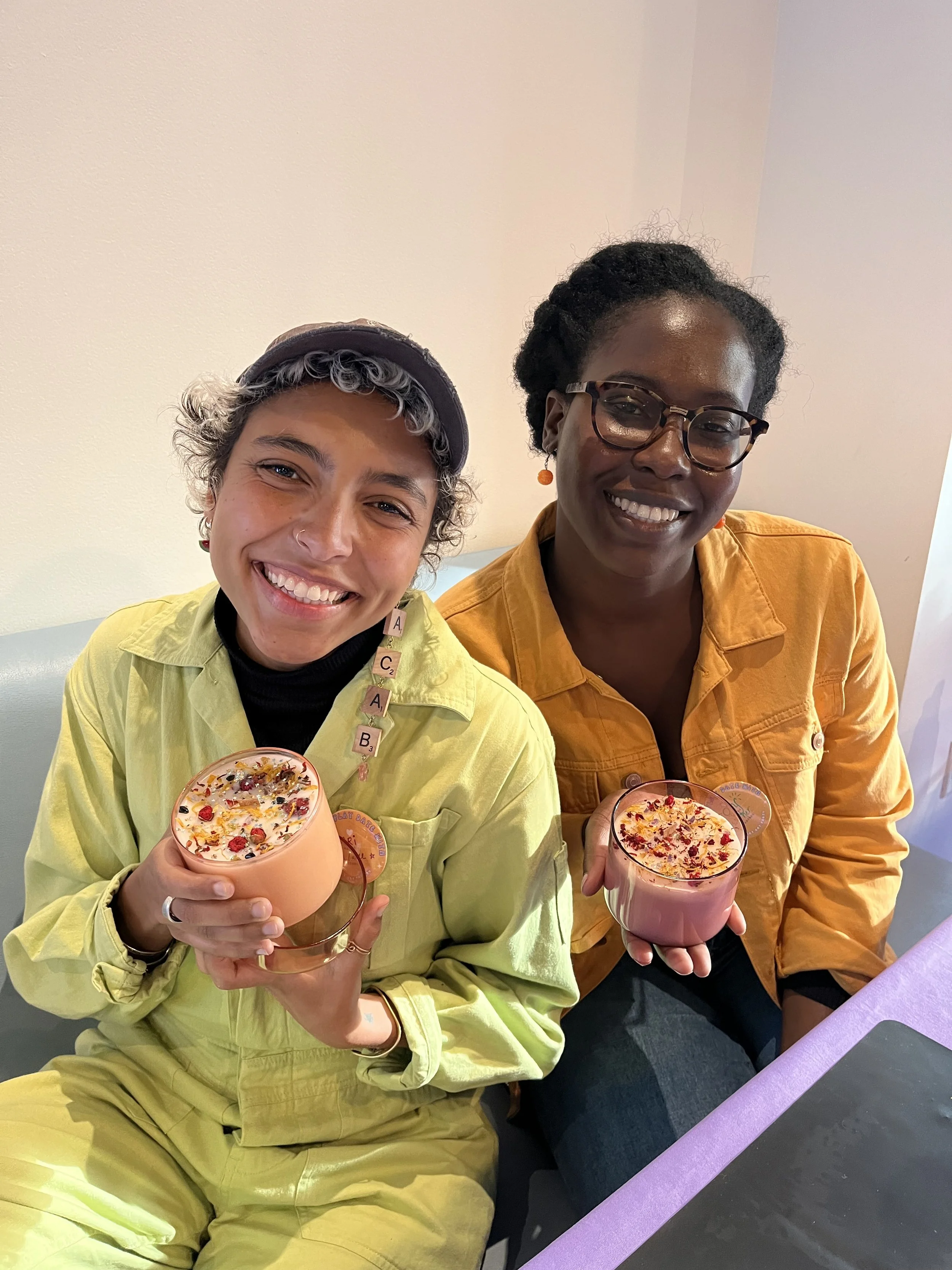 Two people, Black and Latine, holding handmade 12 ounce luxury candles and smiling